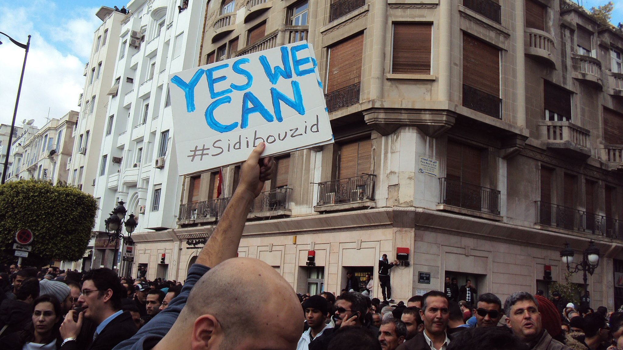 L'image montre une foule rassemblée dans une ville, probablement lors d'une manifestation. Au centre, une personne tient une pancarte avec le message "YES WE CAN" et le hashtag "#Sidibouzid". Les bâtiments en arrière-plan suggèrent un environnement urbain. L'atmosphère semble être celle d'un mouvement populaire, avec des participants engagés et déterminés.