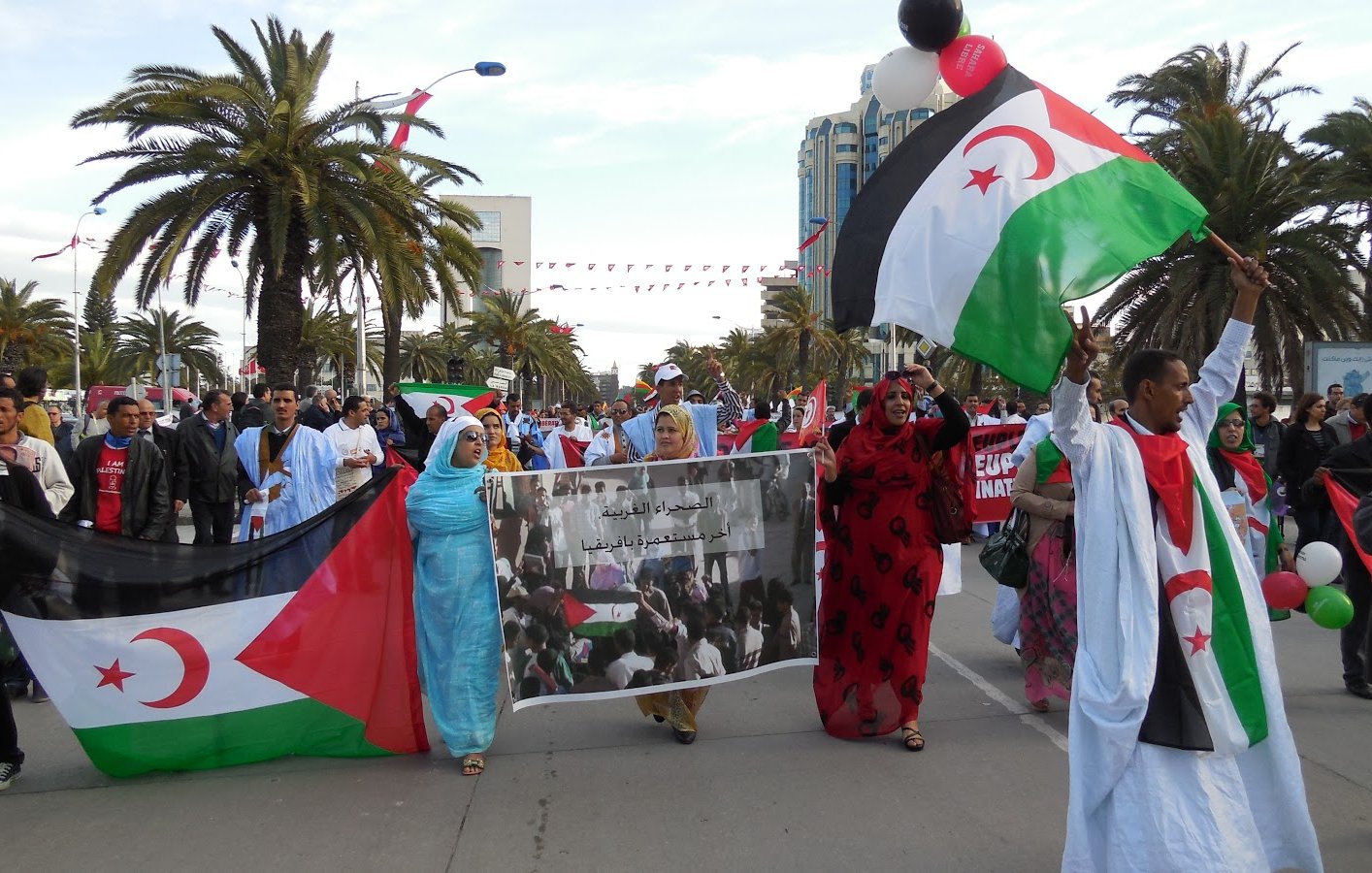 L'image montre une manifestation avec des personnes marchant dans une rue, portant des drapeaux représentant le Sahara occidental. Il y a une atmosphère festive, avec des participants qui tiennent des banderoles et des ballons. Des palmiers se dressent en arrière-plan, ajoutant à l'ambiance. Les manifestants semblent exprimer leurs revendications et leur solidarité.