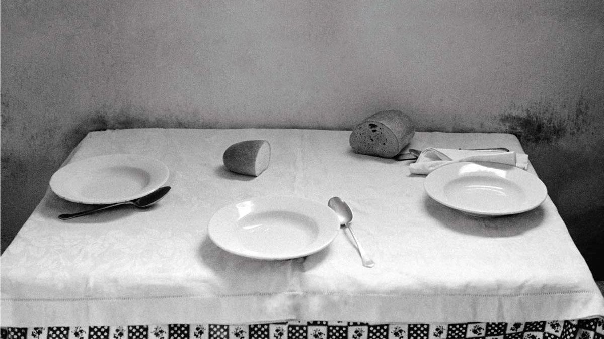 L'image montre une table couverte d'une nappe blanche, où se trouvent deux assiettes blanches, une cuillère et un couteau. Un petit pain est positionné sur un côté et un verre est renversé au centre de la table. L'ambiance générale est sobre et monochrome, ce qui donne une impression de simplicité et de calme. Les murs derrière la table sont légèrement ombragés, ajoutant un effet de profondeur à la composition.