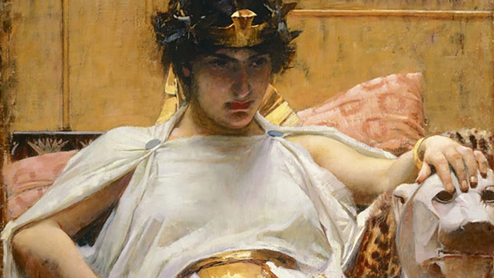 L'image représente une femme assise sur un siège orné, drapée dans une tunique blanche et une écharpe dorée. Elle porte une couronne et a les cheveux sombres, avec un regard pensif et sérieux. À côté d'elle, on aperçoit un lion stylisé sur le siège. L'arrière-plan est décoré de motifs égyptiens, avec des couleurs chaudes et une ambiance luxueuse. La composition évoque une atmosphère royale et mystique.