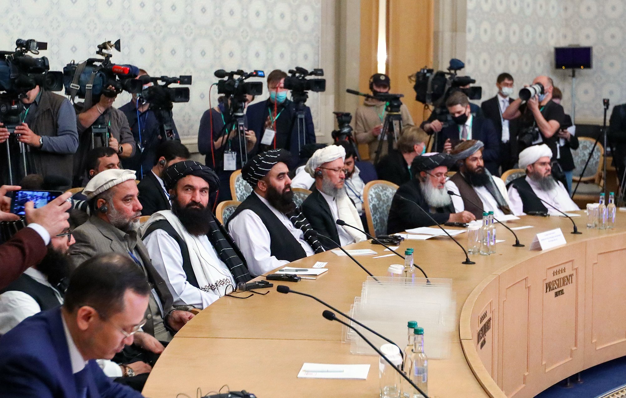 L'image montre une réunion importante avec plusieurs hommes assis en rangée derrière une table. Ils portent des vêtements traditionnels et semblent être des membres d'un groupe politique ou de dirigeants. En arrière-plan, il y a de nombreux journalistes et caméras, suggérant que l'événement est médiatisé. La salle est bien éclairée, avec une décoration neutre et un air d'importance formelle.