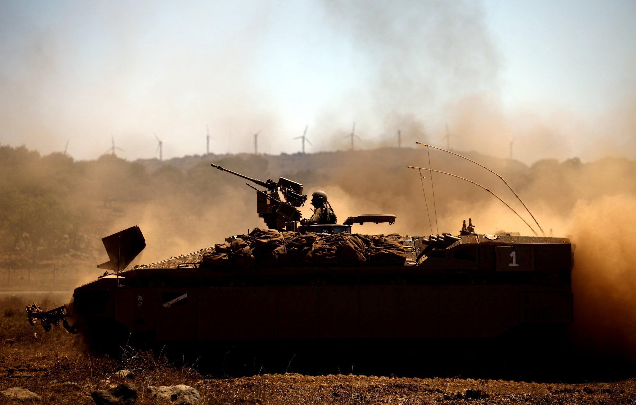 L'image montre un véhicule militaire, probablement un blindé, en action dans un paysage poussiéreux. Un soldat est installé sur le toit du véhicule, armé et prêt. En arrière-plan, on peut apercevoir des éoliennes qui se dressent contre un ciel clair. La scène évoque une ambiance de tension et de préparation militaire, tandis que la poussière soulevée par le mouvement ajoute une dynamique à l'image.