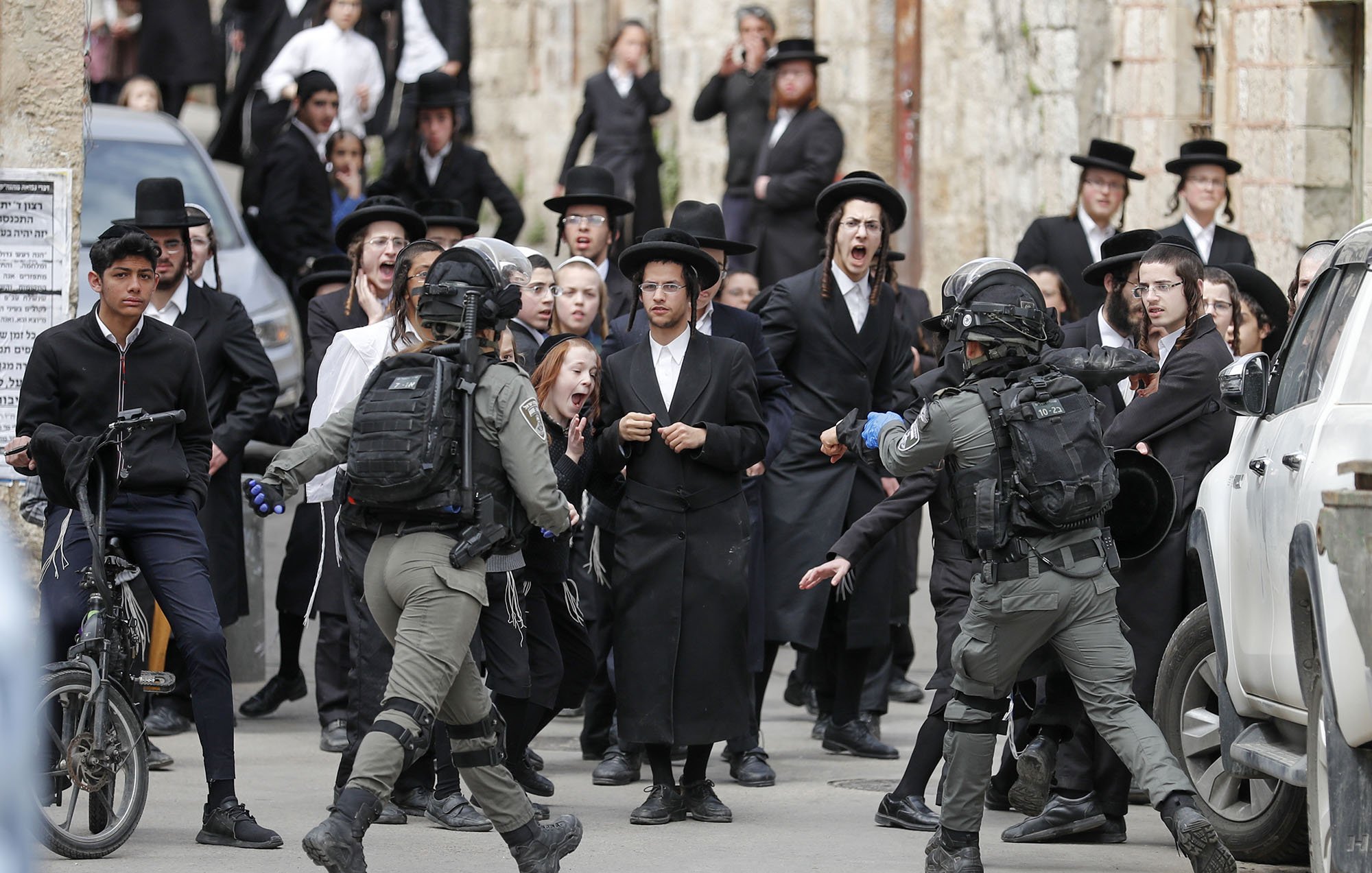 L'image montre une scène animée dans une rue, où un groupe de personnes, principalement habillées en noir, semble manifester ou réagir à une situation tendue. On peut voir des hommes en habits traditionnels, portant des chapeaux noirs, ainsi que des adolescents sur des bicycles. Au centre, des membres des forces de l'ordre, en tenue tactique, interagissent avec la foule. Les expressions faciales des personnes dans la foule varient entre la surprise, l'inquiétude et l'agitation, créant une atmosphère de tensions. Les bâtiments en arrière-plan évoquent un contexte urbain, probablement dans une région où se concentrent des communautés religieuses.