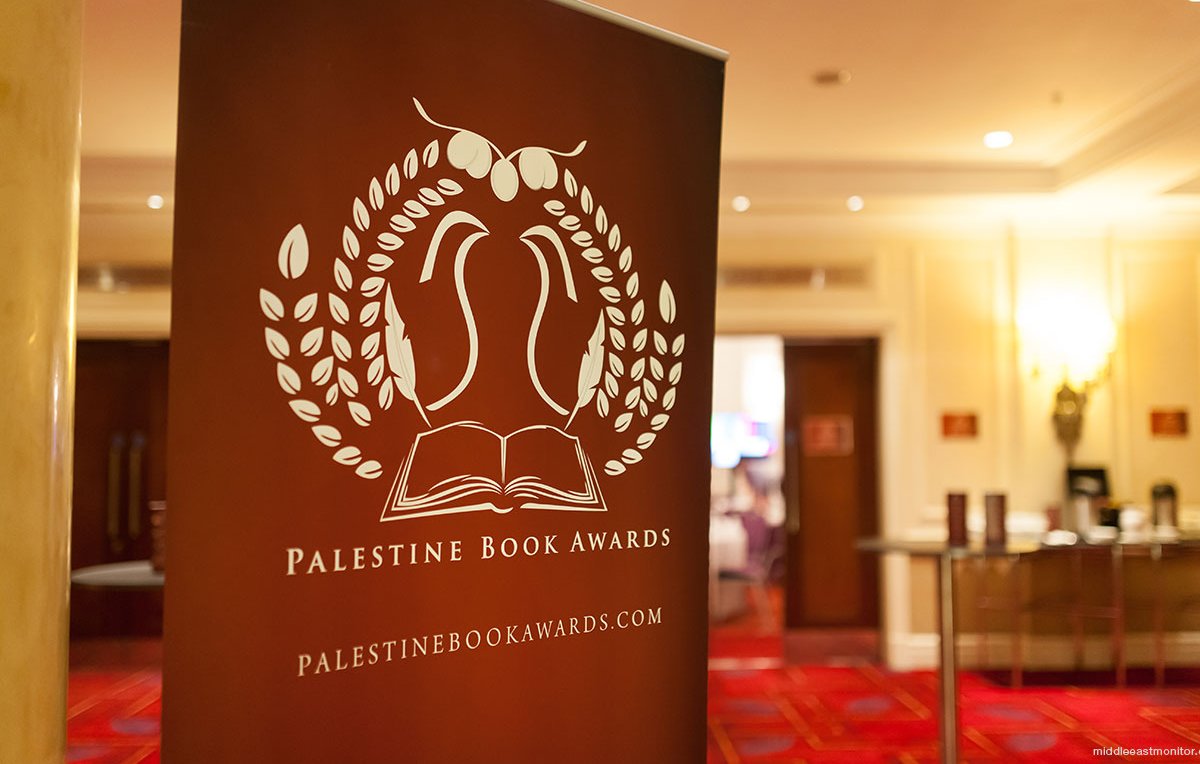 L'image représente un panneau de présentation pour les Palestine Book Awards. Ce panneau arbore un design avec des éléments graphiques, incluant une illustration d'un livre ouvert entouré de feuilles. Le texte indique "Palestine Book Awards" ainsi que le site web associé. L'arrière-plan semble être une salle de réception, avec un sol tapissé et des éclairages doux.