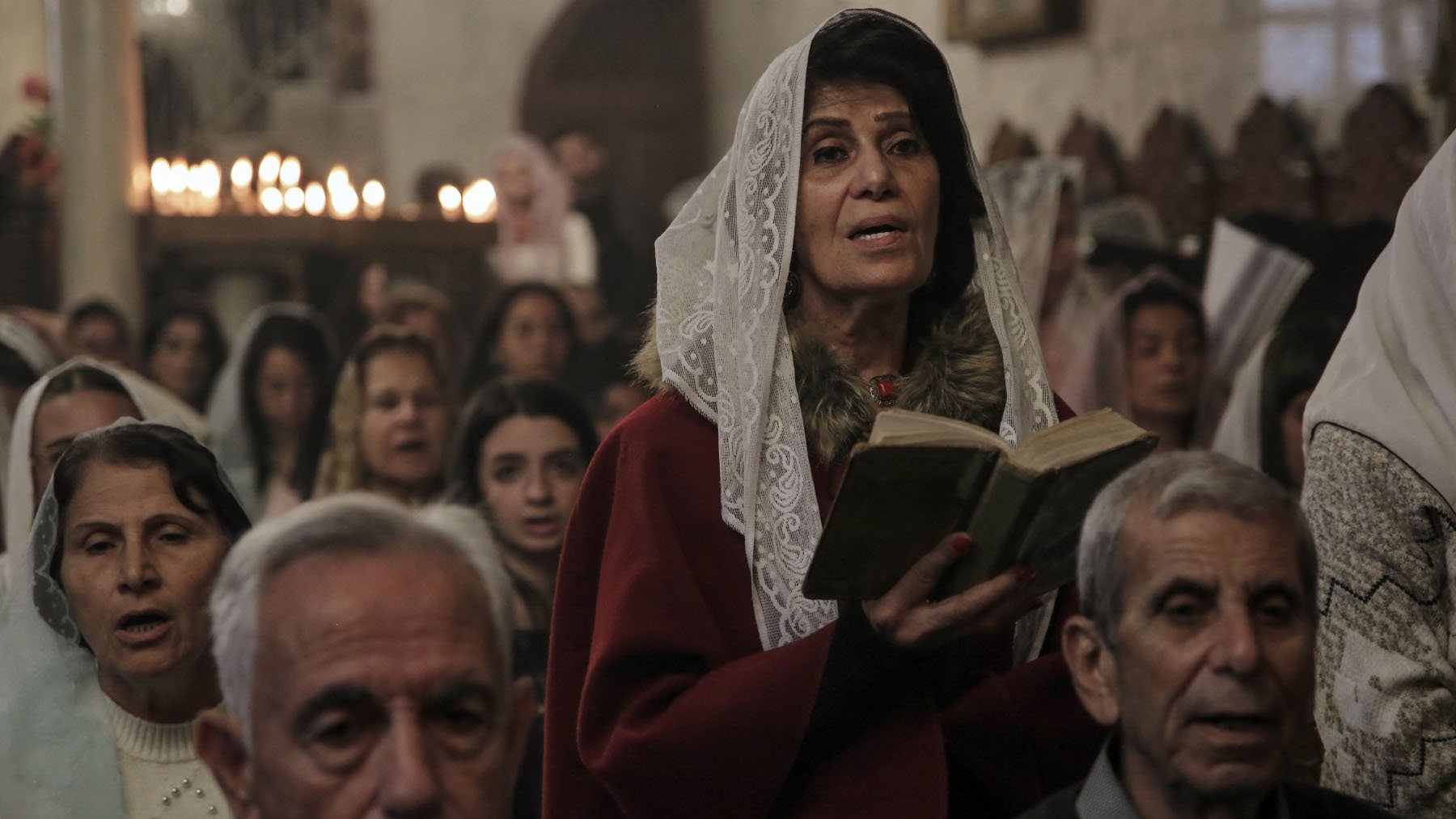 L'image montre un groupe de personnes rassemblées dans un lieu de culte. Au premier plan, une femme portant un vêtement rouge et un voile lit un livre, probablement un texte religieux. En arrière-plan, d'autres personnes, dont certaines semblent chanter ou prier, sont visibles. L'atmosphère semble solennelle, éclairée par des bougies qui créent une ambiance chaleureuse. Les visages des participants expriment une concentration et une dévotion.