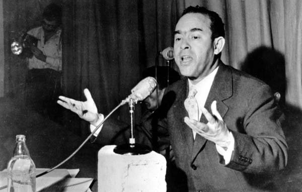 L'image montre un homme en train de parler dans un micro, probablement lors d'une conférence ou d'une réunion. Il a l'air passionné et utilise ses mains pour exprimer ses idées. À l'arrière-plan, on peut voir une personne qui semble être en train de prendre des photos. La scène dégage une atmosphère de discours public ou de débat. L'homme porte un costume et une cravate, ce qui suggère un contexte formel.