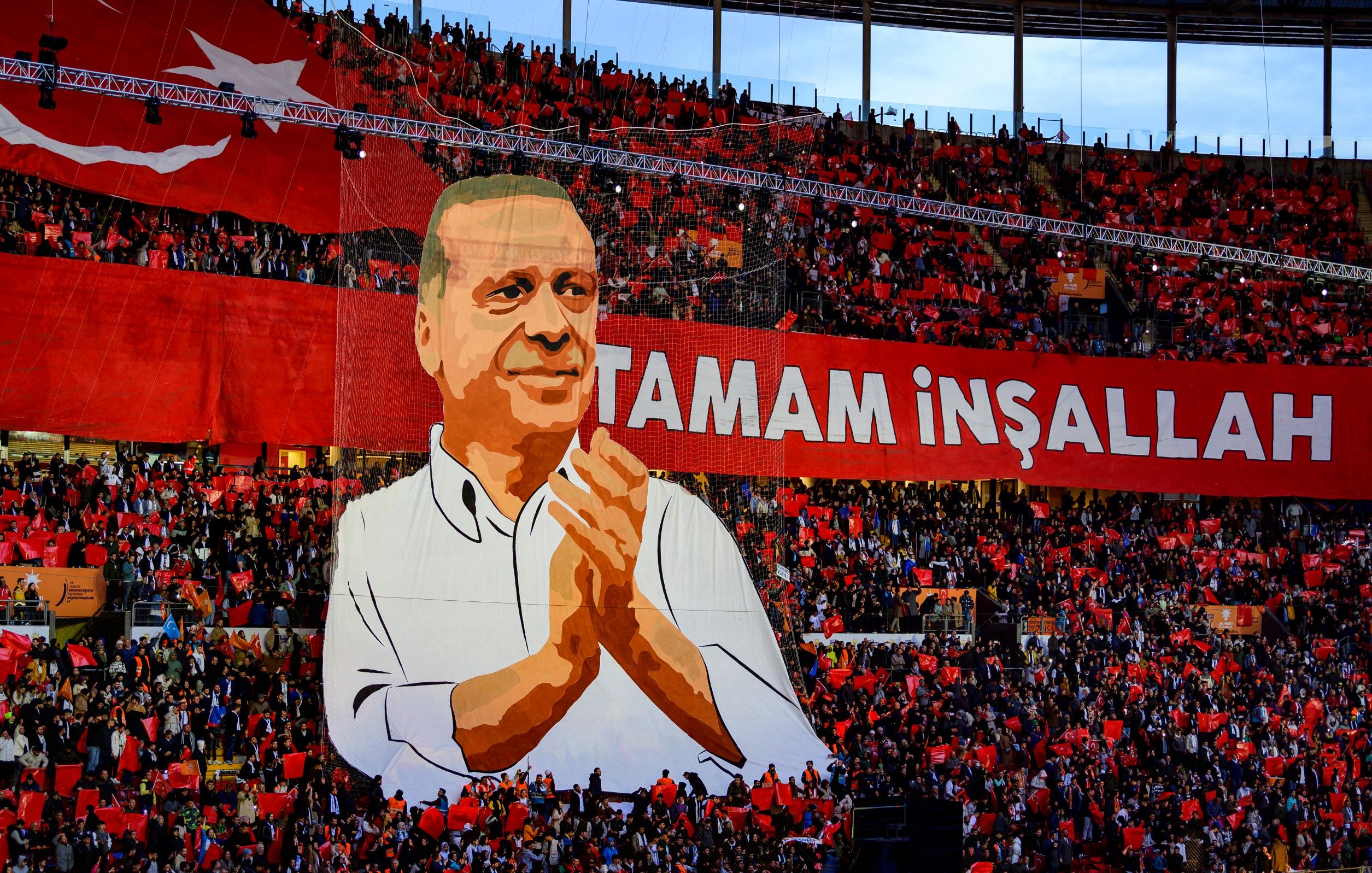 L'image montre une grande scène dans un stade rempli de supporters. Au centre, il y a une immense illustration d'un homme qui semble être un leader ou un personnage politique, probablement Erdoğan, qui applaudit. En arrière-plan, une banderole rouge avec des inscriptions en lettres blanches affiche "TAMAM İNŞALLAH". Les spectateurs tiennent des drapeaux rouges, créant une atmosphère colorée et festive. L'ensemble évoque une manifestation de soutien, probablement lors d'un événement politique.