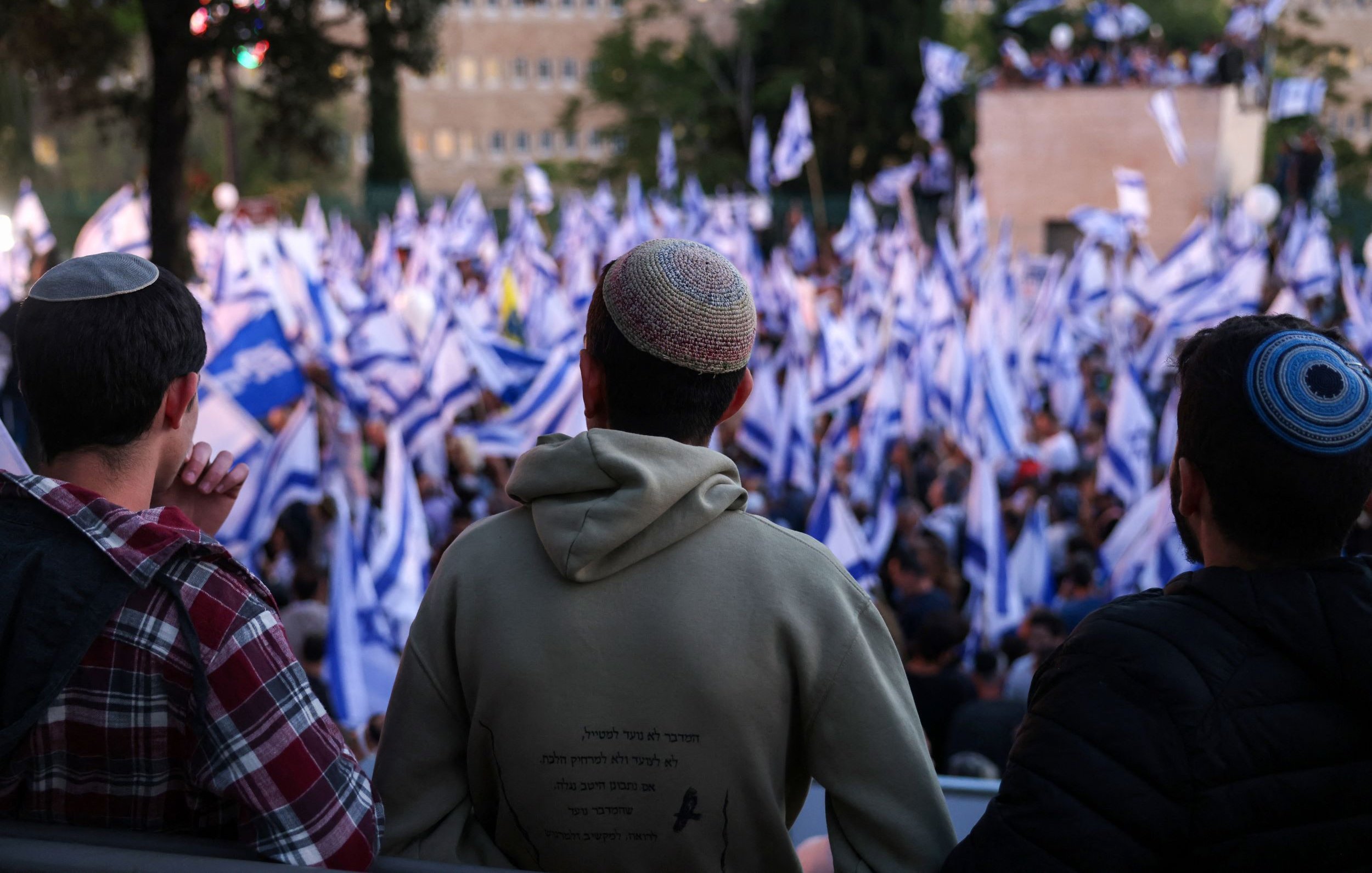 L'image montre un groupe de personnes vu de dos, regardant une manifestation ou un rassemblement. Ils portent des kippas. Au premier plan, on observe un homme en sweat à capuche, tandis que les autres sont légèrement flous. En arrière-plan, une foule dense agite des drapeaux, probablement israéliens, créant une ambiance dynamique. La scène semble se dérouler à l'extérieur, avec des arbres et des bâtiments visibles au loin.