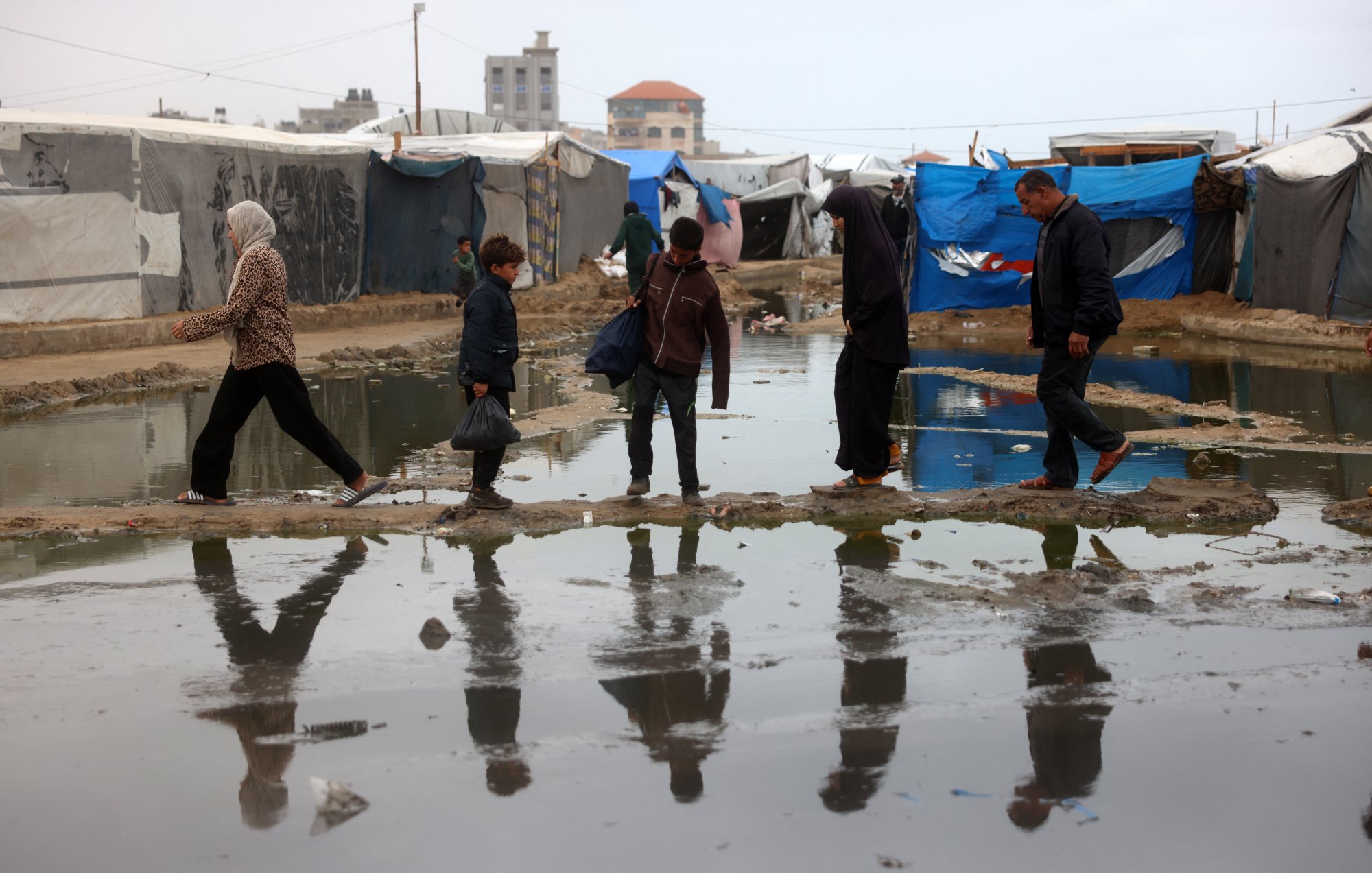 L'image montre des personnes marchant à travers un camp de tentes, avec des surfaces détrempées et des flaques d'eau. On peut voir une femme en hijab, un enfant, et plusieurs adultes, tous avançant sur des passages surélevés ou des morceaux de terrain au-dessus de l'eau. L'environnement semble difficile, et les refuges sont constitués de bâches et de tissus. Les reflets des marcheurs se voient dans les flaques, ajoutant une dimension visuelle à la scène.
