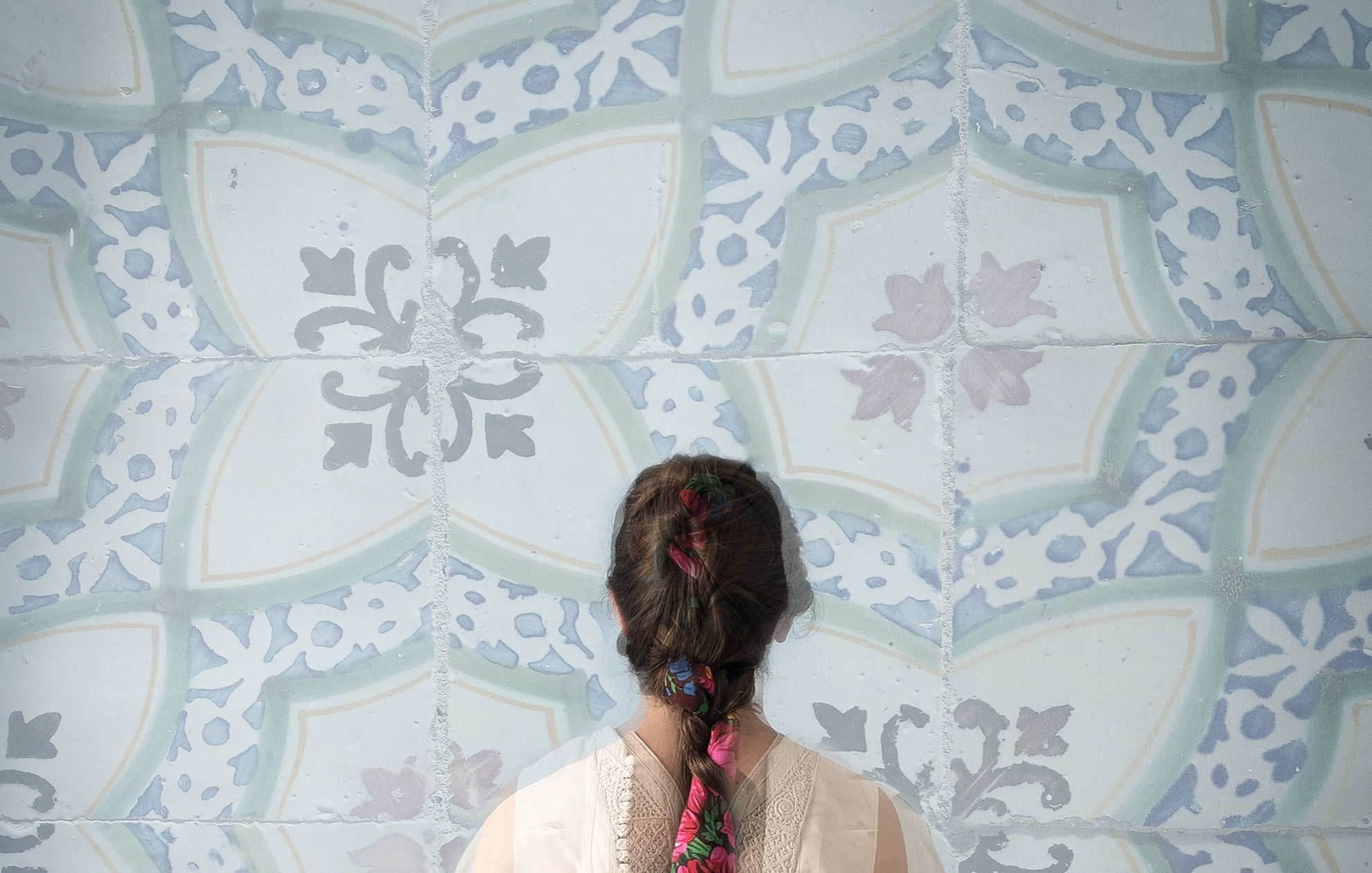 Cette image montre une personne de dos, portant un vêtement léger et délicat. Sa longue tresse, ornée de fleurs colorées, est visible et se détache sur un fond de motifs bleu clair. Les motifs sont ornés de formes géométriques et florales, créant une ambiance apaisante et artistique. L'ensemble évoque une atmosphère de calme et de beauté.