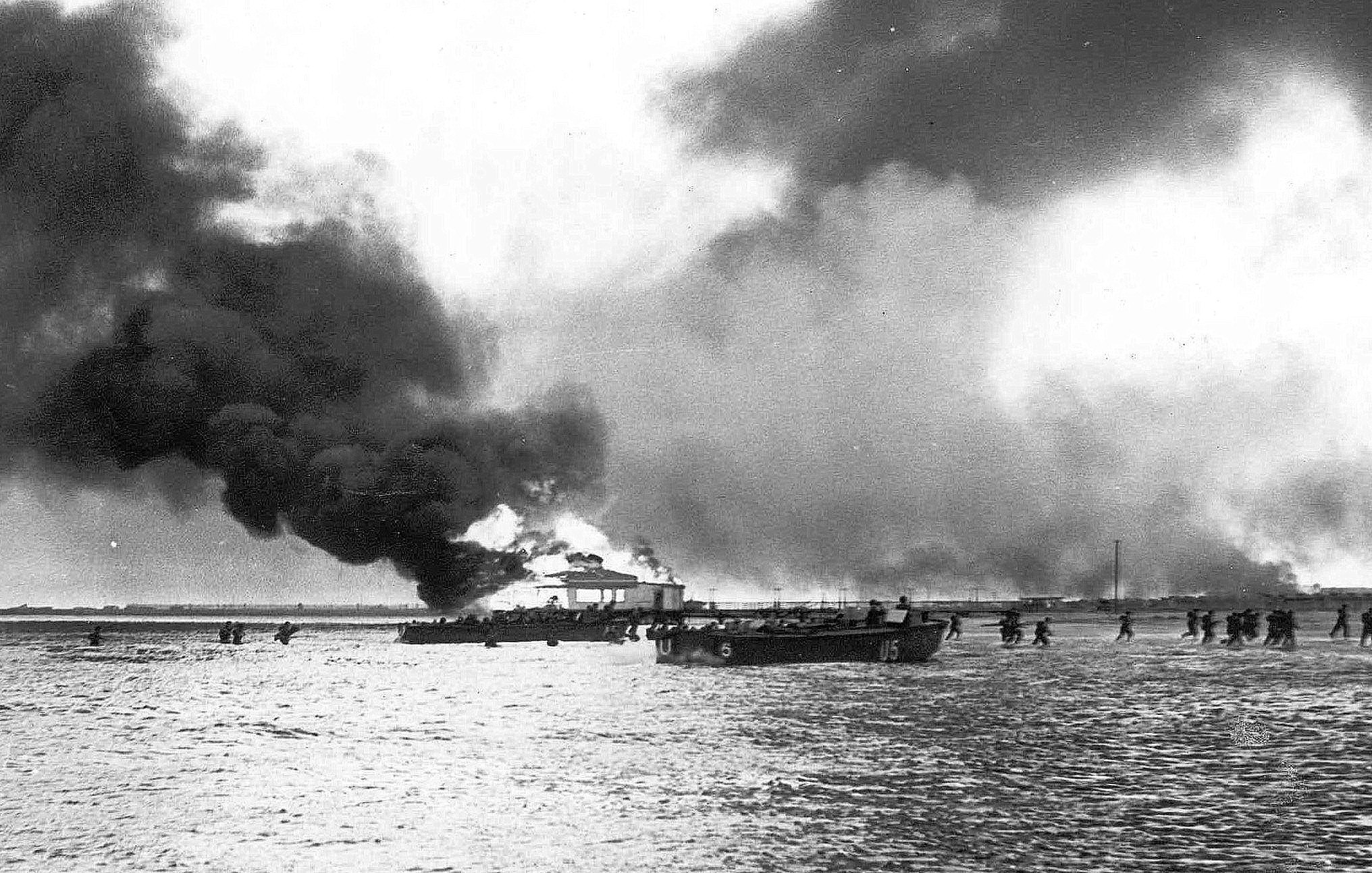 L'image montre une scène sombre et dramatique, probablement liée à un événement de guerre ou de désastre. On peut apercevoir une grande quantité de fumée noire s'élevant dans le ciel, suggérant qu'un bâtiment ou un navire est en train de brûler. Au premier plan, il y a un bateau sur l'eau, et au loin, plusieurs personnes semblent marcher le long de la rive, potentiellement évacuant une zone touchée par le feu ou un conflit. L'atmosphère générale est sombre et chaotique, avec des éléments de destruction visibles.