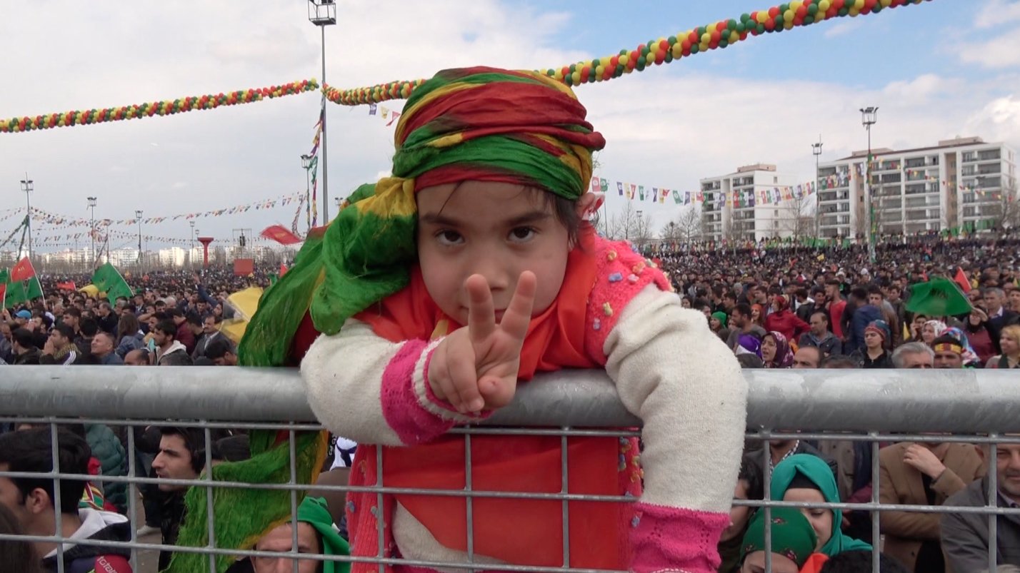 L'image montre une jeune fille participant à un grand rassemblement. Elle est vêtue de vêtements colorés, portant un foulard aux teintes vives. La fillette fait un signe de paix avec ses doigts, pendant qu'elle se penche sur une barrière. En arrière-plan, une grande foule est présente, témoignant d'un événement festif ou d'une manifestation populaire, avec des décorations colorées suspendues au-dessus. L'ambiance semble joyeuse et engagée.