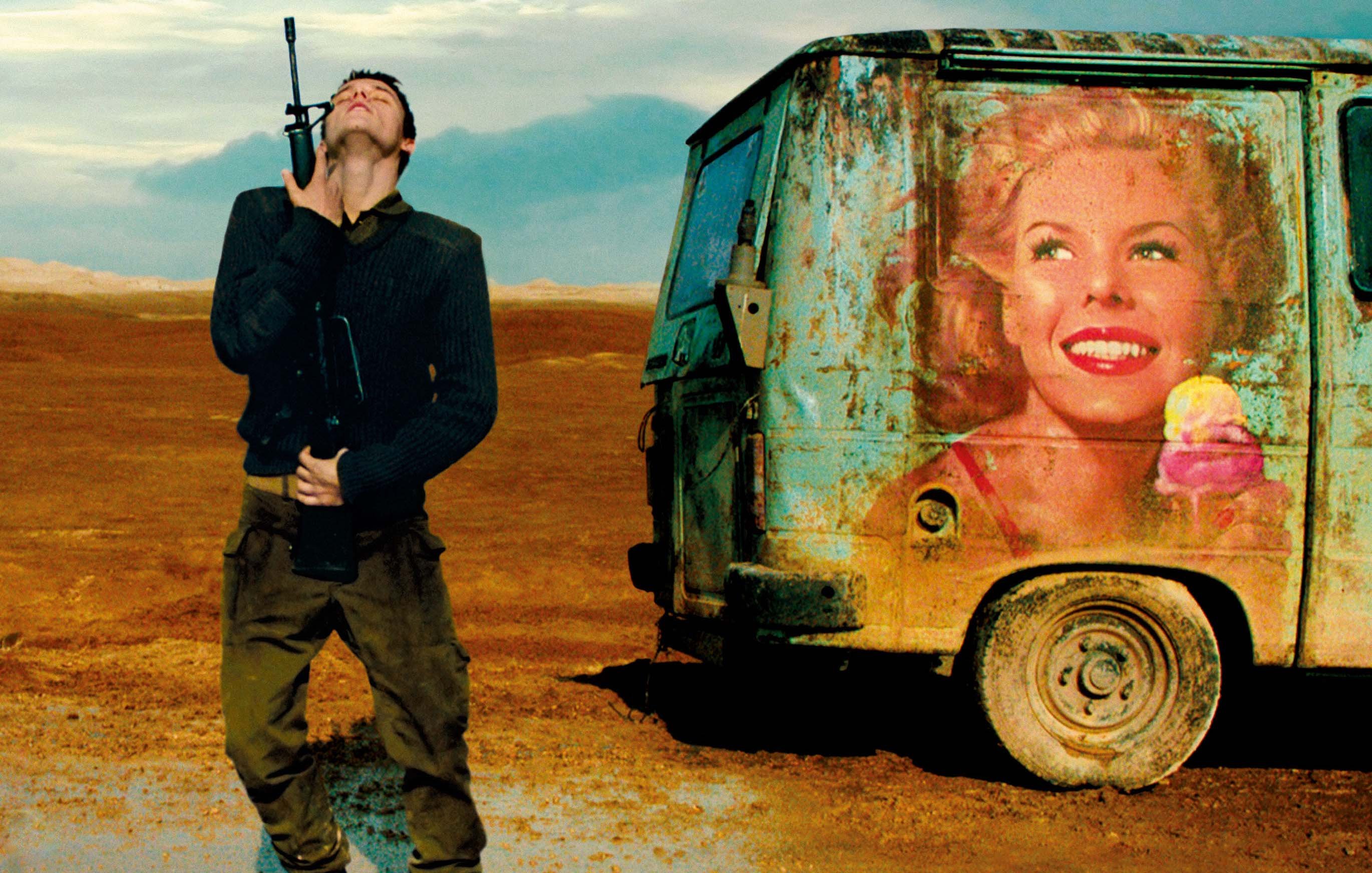 L'image montre un homme debout dans un paysage désertique, avec une expression qui semble mélancolique ou contemplative. Il porte une arme à feu à la main et est habillé de manière militaire. À côté de lui se trouve un vieux camion, sur lequel est peint un portrait coloré d'une femme souriante tenant une glace. Le contraste entre l'aspect rustique du véhicule et l'image joyeuse de la femme crée une atmosphère intrigante, mêlant des éléments de décor et d'émotion. Le ciel, quant à lui, est nuageux, ajoutant une dimension dramatique à la scène.