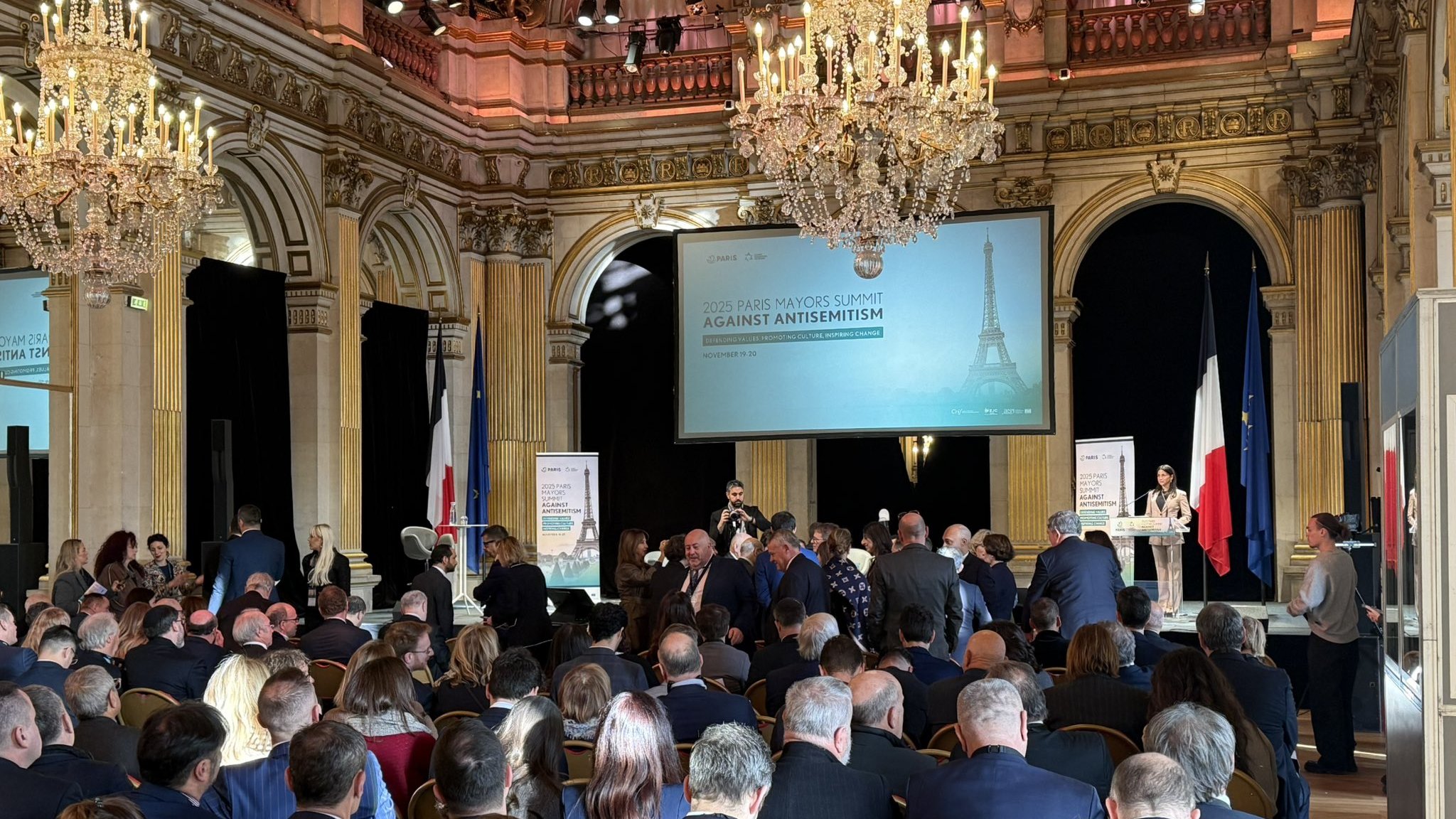 Grand hall avec chandeliers, foule réunie autour d'un discours contre l'antisémitisme.
