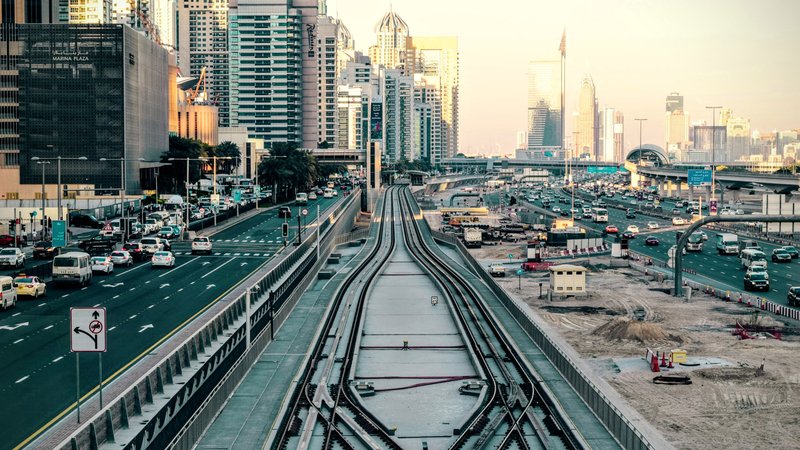 L'image montre une vue urbaine d'une ville moderne. On peut voir des routes animées avec des voitures circulant, ainsi qu'un important réseau de rails de train. À droite, il y a des bâtiments imposants et modernes qui reflètent l'architecture contemporaine. L'atmosphère est lumineuse, suggérant un moment de la journée proche du coucher du soleil avec des lumières douces. Les rails semblent bifurquer, suggérant une intersection, tandis que des travaux de construction ou d'aménagement sont visibles au premier plan.