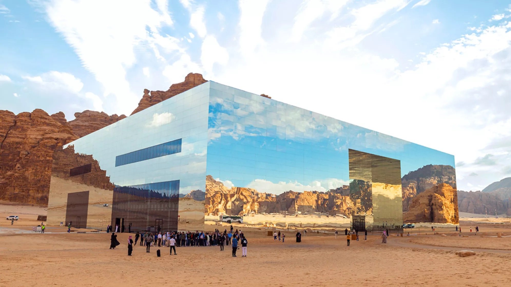 L'image montre un bâtiment moderne et futuriste situé dans un paysage désertique. La structure est entièrement recouverte de miroirs, reflétant le ciel et les montagnes environnantes, ce qui lui permet de se fondre harmoniellement dans le paysage. Au premier plan, un groupe de personnes, vraisemblablement des visiteurs, se regroupe sur le sable. L'atmosphère est paisible, et le ciel est parsemé de nuages, créant un contraste avec les teintes désertiques. L'architecture innovante et les réflexions ajoutent une dimension visuelle fascinante à l'environnement naturel.
