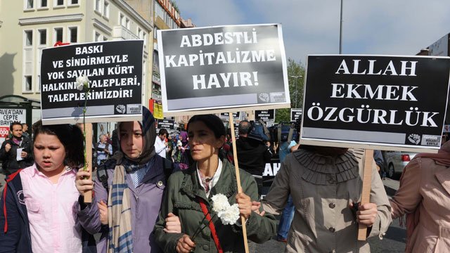L'image montre une manifestation avec des personnes portant des pancartes. Les manifestants semblent exprimer des revendications contre la pression et l'oppression, en mettant en avant des messages en turc. Certaines femmes portent des foulards, suggérant un contexte culturel spécifique. On peut sentir une atmosphère de mobilisation collective.