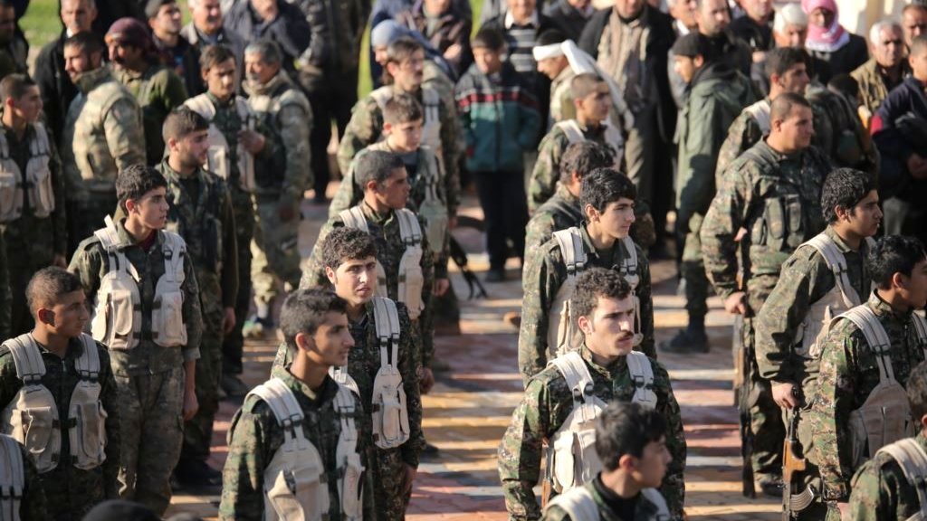 L'image montre un groupe de soldats en formation, portant des uniformes militaires et des équipements. Ils sont alignés, en regardant droit devant eux, et semblent être en train de participer à une cérémonie ou à un événement officiel. En arrière-plan, on peut observer une foule de personnes, probablement des civils, qui les regardent. L'atmosphère semble sérieuse et solennelle.