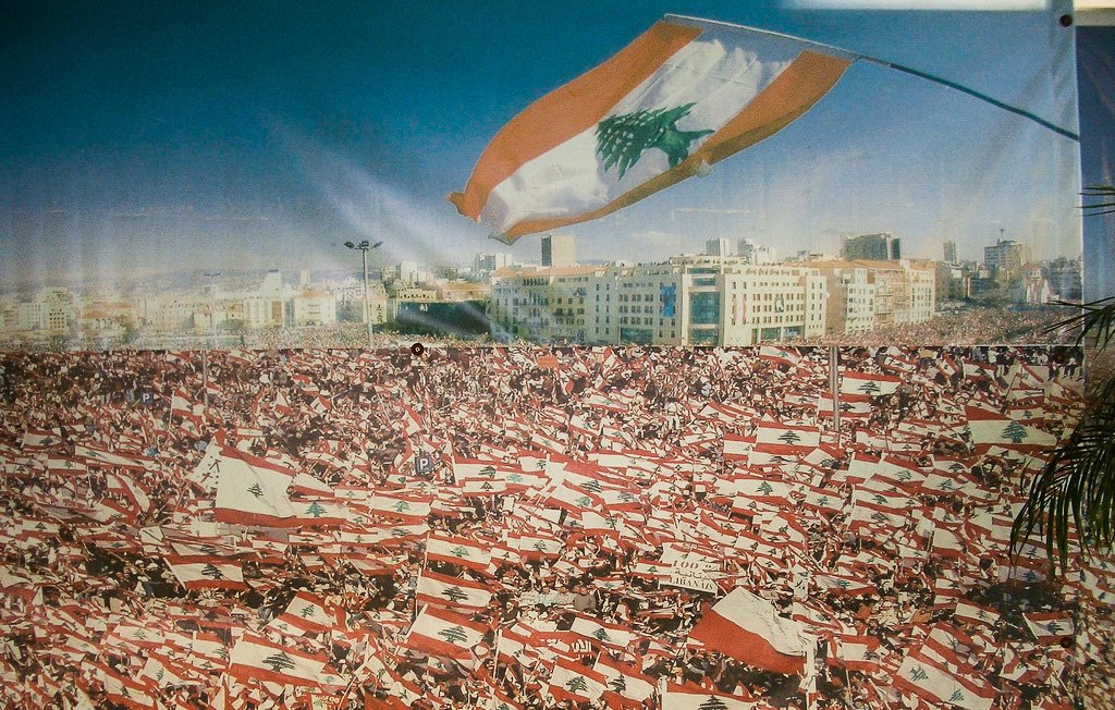 L'image montre une grande foule rassemblée, probablement lors d'une manifestation ou d'un événement public au Liban. La mer de personnes brandit de nombreux drapeaux libanais, créant un effet visuel saisissant. En arrière-plan, on aperçoit une ville moderne, ce qui suggère une ambiance vibrante. Le ciel est dégagé, ajoutant à la luminosité de la scène. Cette image évoque un sentiment de patriotisme et de solidarité parmi les participants.