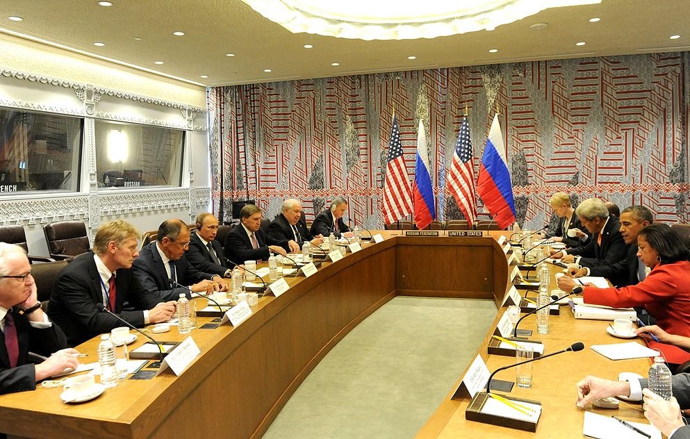 L'image montre une réunion formelle se déroulant dans une salle de conférence. À une longue table, des représentants des États-Unis et de la Russie sont présents, avec des drapeaux des deux pays visibles en arrière-plan. Les participants, qui semblent être des diplomates ou des responsables gouvernementaux, sont engagés dans une discussion sérieuse. La décoration de la salle présente des motifs géométriques sur les murs, et l'ambiance générale est celle d'une négociation ou d'un dialogue diplomatique.