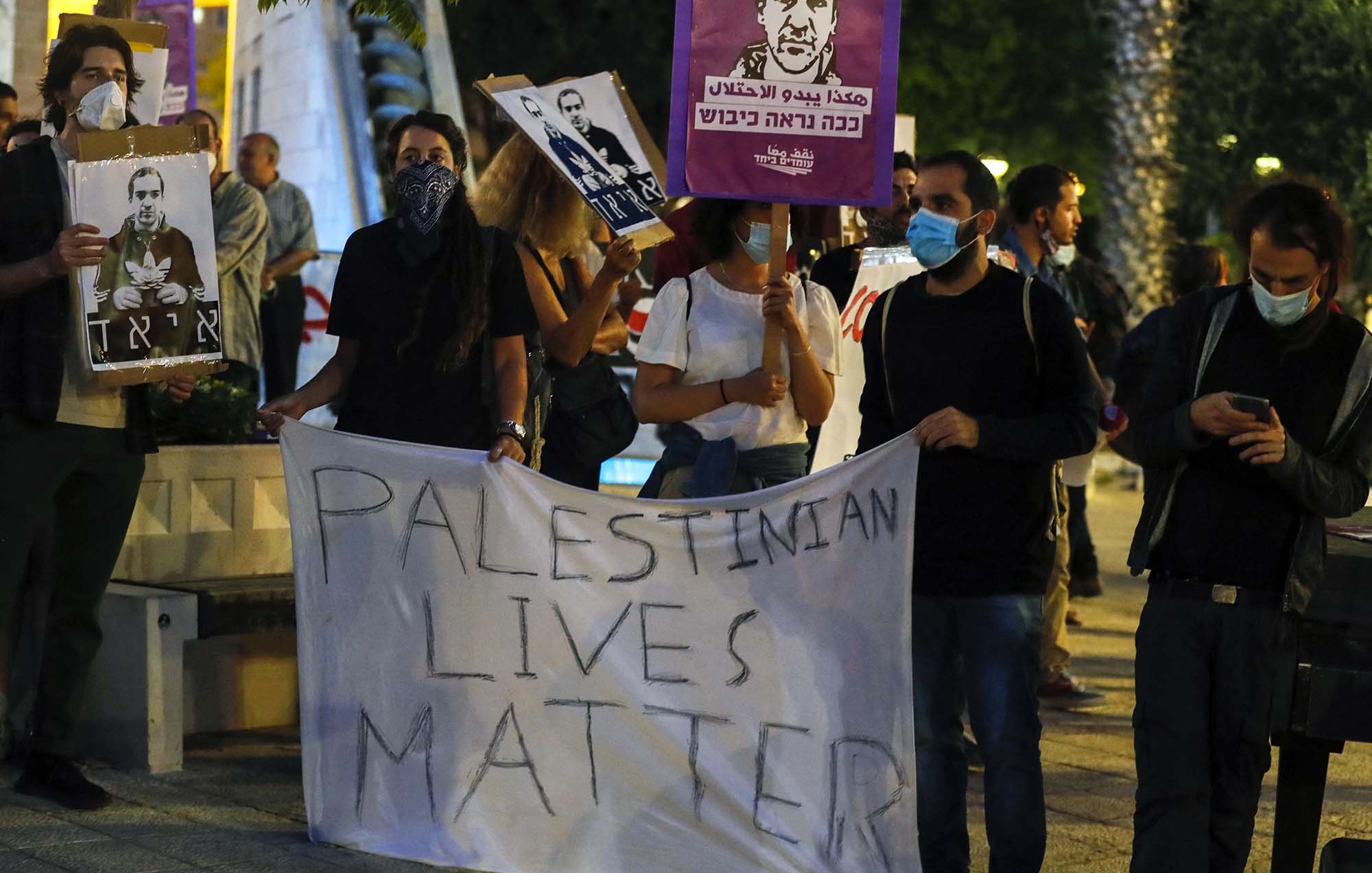 L'image montre un groupe de personnes manifestant. Elles tiennent des panneaux et une grande banderole sur laquelle est écrit "PALESTINIAN LIVES MATTER". Les participants portent des masques, et certains affichent des portraits d'individus. L'ambiance semble être celle d'une manifestation pour les droits des Palestiniens. Le décor urbain, avec des arbres et des lumières en arrière-plan, ajoute à l'atmosphère de l'événement.