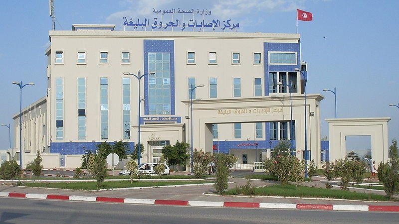 L'image montre un bâtiment moderne, probablement un centre de santé ou une institution gouvernementale. La façade est de couleur claire avec des éléments architecturaux en bleu. On y voit des fenêtres de différentes tailles et une grande enseigne en arabe et en français. Un drapeau tunisien flotte sur le toit, ce qui indique que le bâtiment est situé en Tunisie. L'environnement semble soigné, avec un espace urbain bien aménagé autour du bâtiment.