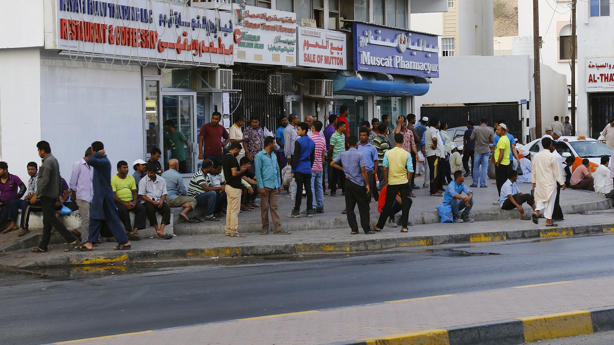 L'image montre une scène urbaine animée, avec un groupe de personnes rassemblées devant un bâtiment. On peut voir des hommes de différentes tenues discutant, assis ou se tenant debout sur le trottoir. Le bâtiment porte des enseignes de restaurants et de pharmacie, indiquant une ambiance commerçante. La route est visible au premier plan, avec une bande jaune délimitant la chaussée. L'atmosphère semble vivante, typique des zones urbaines fréquentées.