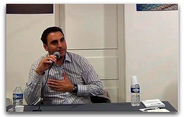 L'image montre un homme assis à une table, tenant un microphone. Il porte une chemise à rayures et semble s'exprimer avec conviction. Sur la table, il y a une bouteille d'eau et un gobelet, ainsi que des documents visibles. L'arrière-plan montre des affiches ou des œuvres, mais les détails ne sont pas clairement discernables.