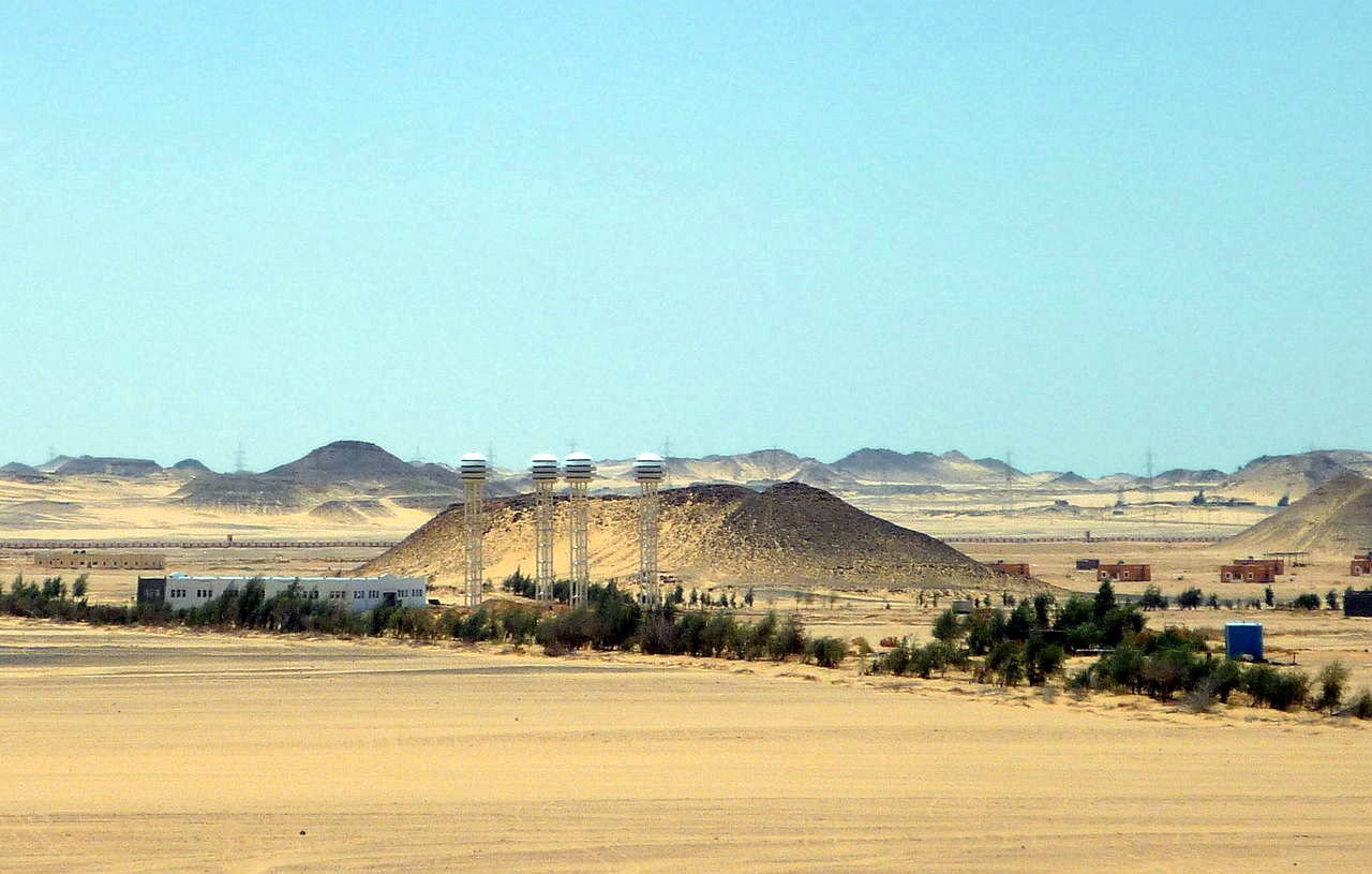 L'image montre un paysage désertique vaste et aride, avec des dunes de sable et des collines. Au centre, on peut distinguer quelques bâtiments qui semblent être des installations humaines, entourés de végétation telle que des arbres ou des buissons. À l'arrière-plan, des collines se découpent contre le ciel, tandis qu'une lumière vive souligne l'immensité du paysage. L'atmosphère semble calme et isolée, typique des régions désertiques.