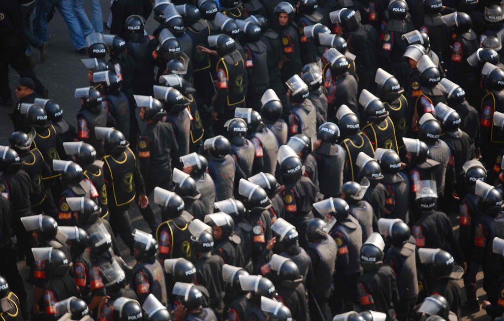 L'image montre un grand groupe de policiers en uniforme, portant des casques et des protections. Ils sont disposés de manière organisée et semblent être en formation, probablement lors d'une manifestation ou d'un événement de sécurité. Les uniformes sont généralement noirs avec des éléments de couleur, et on peut voir des panneaux de protection en plastique. L'atmosphère semble intense et sérieuse, reflétant une situation de gestion de la foule.