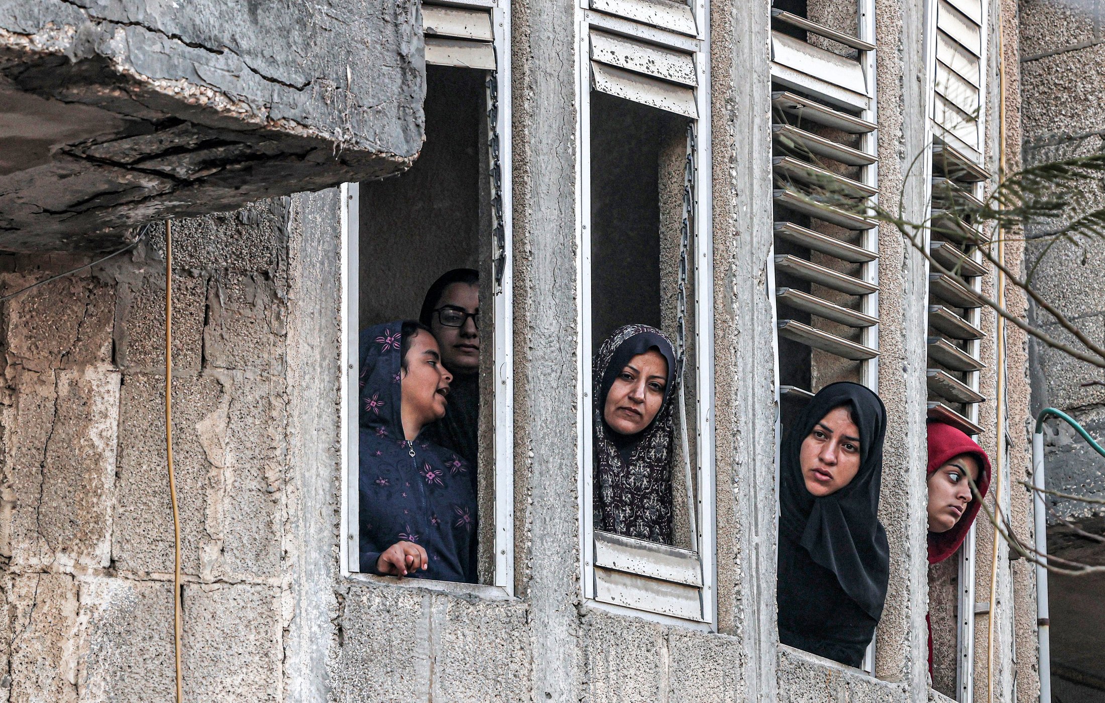 L'image montre plusieurs femmes regardant par des fenêtres d'un bâtiment. Elles semblent attentives et curieuses, certaines portant des hijabs. Le mur extérieur du bâtiment est en béton, et on peut apercevoir des fenêtres aux volets cassés. L'ambiance paraît chargée d'émotions, mais on ne peut pas déterminer le contexte exact de la scène.
