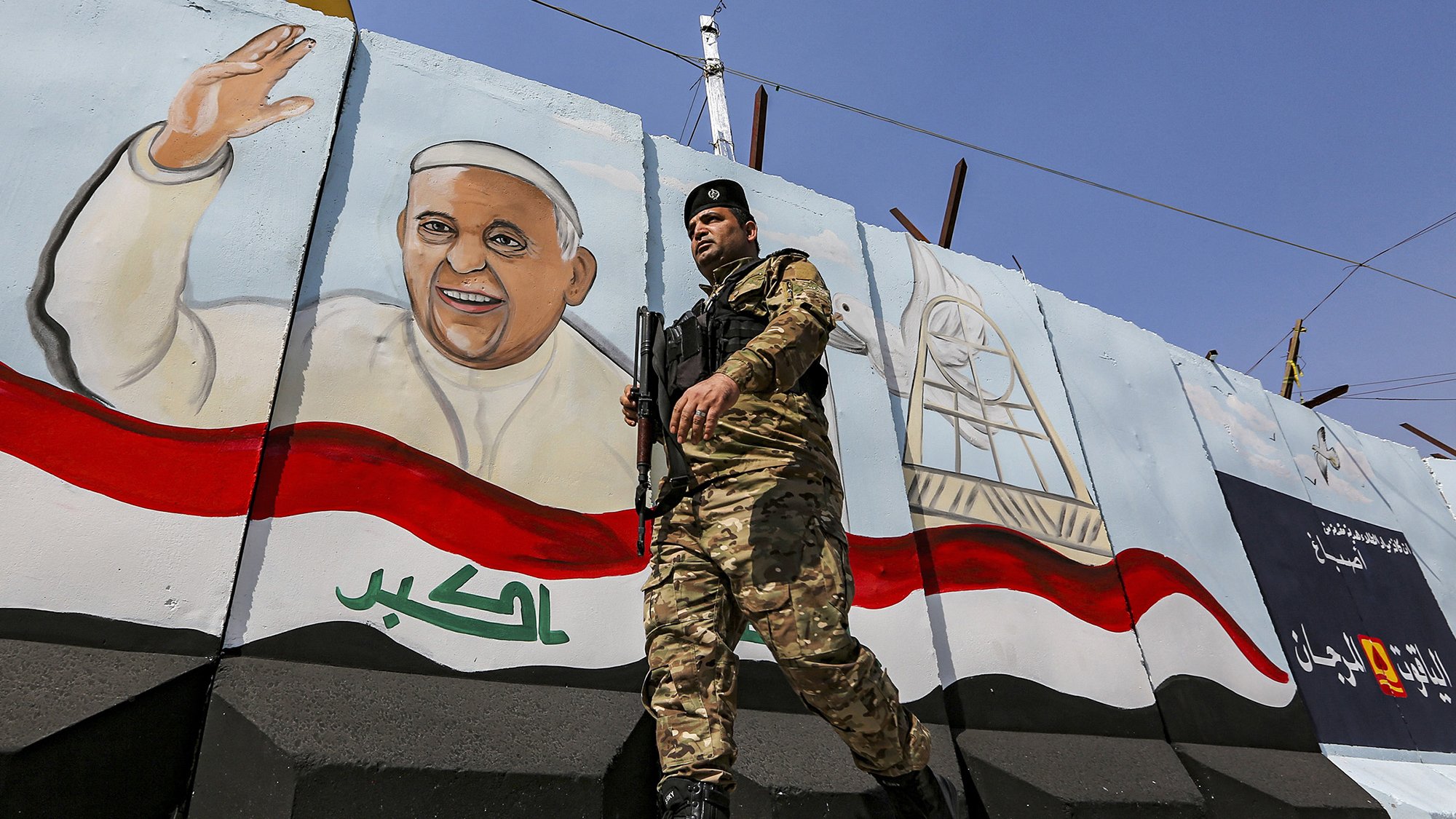 L'image montre un soldat en uniforme, armé et en marche, devant un mur peint. Sur le mur, il y a une fresque représentant un homme souriant et levant la main, probablement symbolisant une figure importante ou un leader. Le fond de la fresque inclut des éléments décoratifs et des couleurs qui évoquent le drapeau irakien. L'environnement semble être une zone urbaine, marquée par des installations de sécurité.
