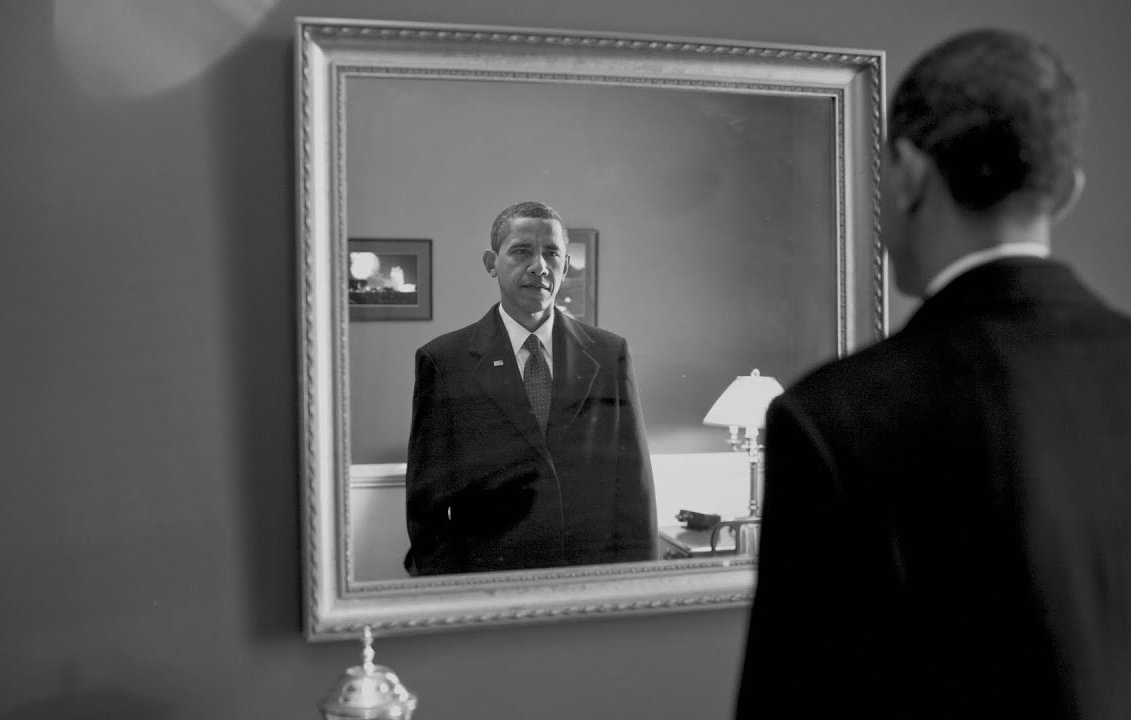 L'image montre un homme en costume noir se tenant devant un miroir. Son reflet dans le miroir affiche une expression sérieuse. L'environnement semble sobre, avec des éléments de décoration tels qu'une lampe et un cadre au mur. La scène est présentée en noir et blanc, ajoutant une atmosphère mélancolique et introspective à l'image.