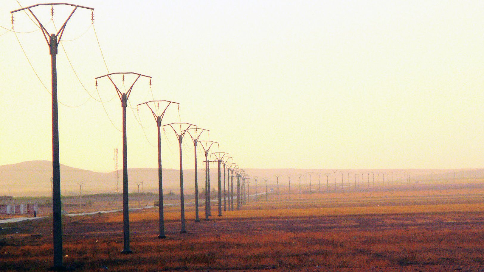 L'image montre une perspective sur un paysage de campagne, dominé par des pylônes électriques s'étendant sur une longue distance. Le sol est principalement sec et herbeux, avec des teintes dorées, suggérant la lumière douce d'un lever ou d'un coucher de soleil. En arrière-plan, on peut apercevoir des collines ou des montagnes floues, contribuant à une ambiance calme et désertique. L'ensemble évoque un sentiment d'immensité et de tranquillité.