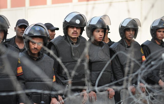L'image montre un groupe de policiers en tenue anti-émeute, portant des casques et des gilets pare-balles. Ils semblent être en formation et regardent attentivement dans une direction, manifestant une posture de vigilance. En arrière-plan, on peut voir des barbelés, ce qui indique un contexte de sécurité renforcée ou de tensions. Les soldats portent des uniformes sombres et sont alignés, ajoutant à l'impression de discipline et de préparation.