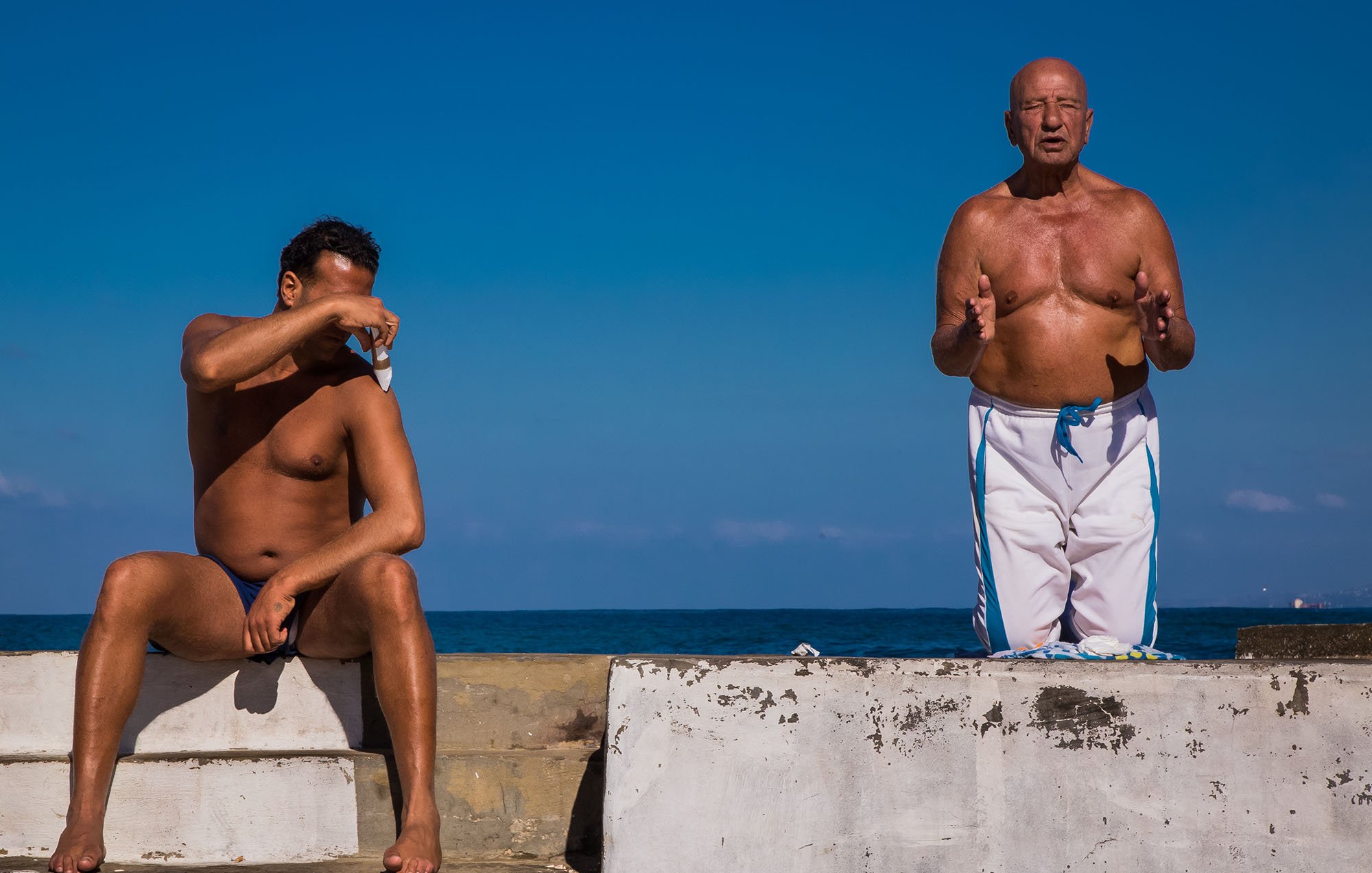L'image montre deux hommes sur une structure en béton au bord de l'eau. L'un des hommes est assis, torse nu, avec les bras croisés sur un genou, semblant protéger ses yeux du soleil. L'autre homme, debout à côté de lui, est également torse nu et semble parler ou gesticuler, comme s'il était en pleine conversation. En arrière-plan, on peut voir un ciel bleu clair et une étendue de mer, créant une atmosphère estivale et décontractée.
