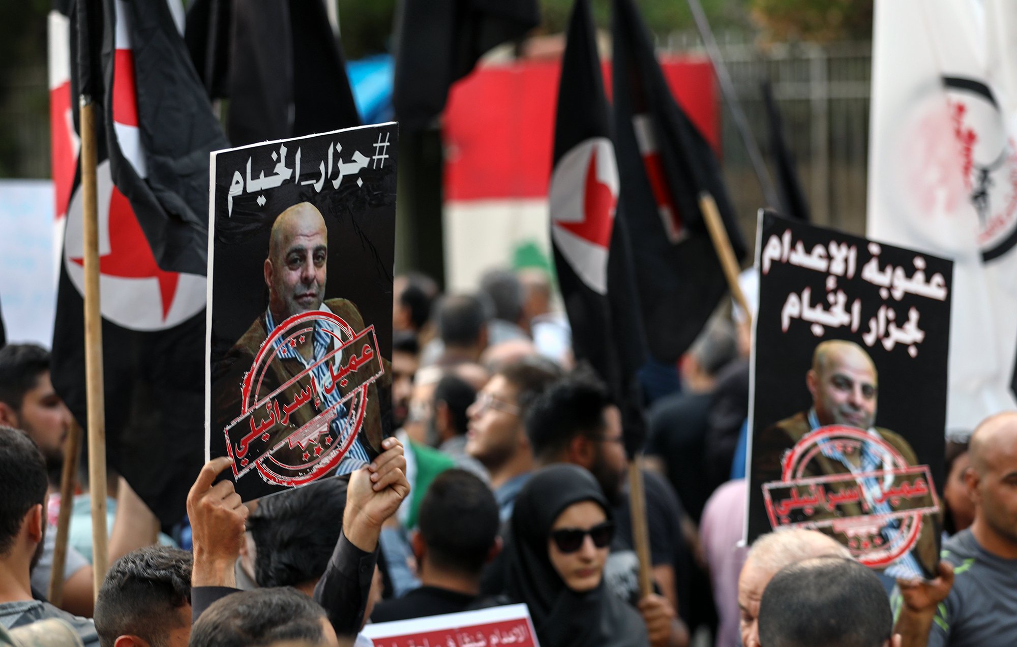 L'image montre une manifestation avec des personnes tenant des pancartes et des drapeaux. Les pancartes contiennent des portraits de personnes et des textes en arabe, évoquant des messages politiques ou sociaux. La foule semble engagée dans une cause, exprimant des émotions fortes. On peut voir une diversité de participants, certains portant des vêtements traditionnels et d'autres plus modernes, ainsi qu'un fond urbain suggérant un rassemblement public.