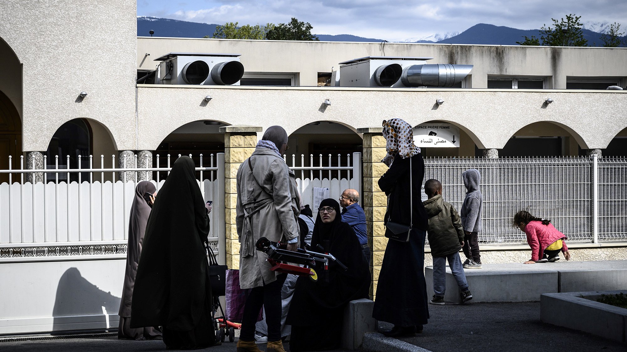 L'image montre un groupe de personnes à l'extérieur d'un bâtiment. Certaines femmes portent des vêtements longs et des foulards, tandis que d'autres semblent habillées plus décontractées. On aperçoit également des enfants jouant dans l'arrière-plan. Le ciel est partiellement nuageux et des montagnes se dessinent au loin. L'ambiance semble conviviale, avec des échanges entre les personnes présentes.