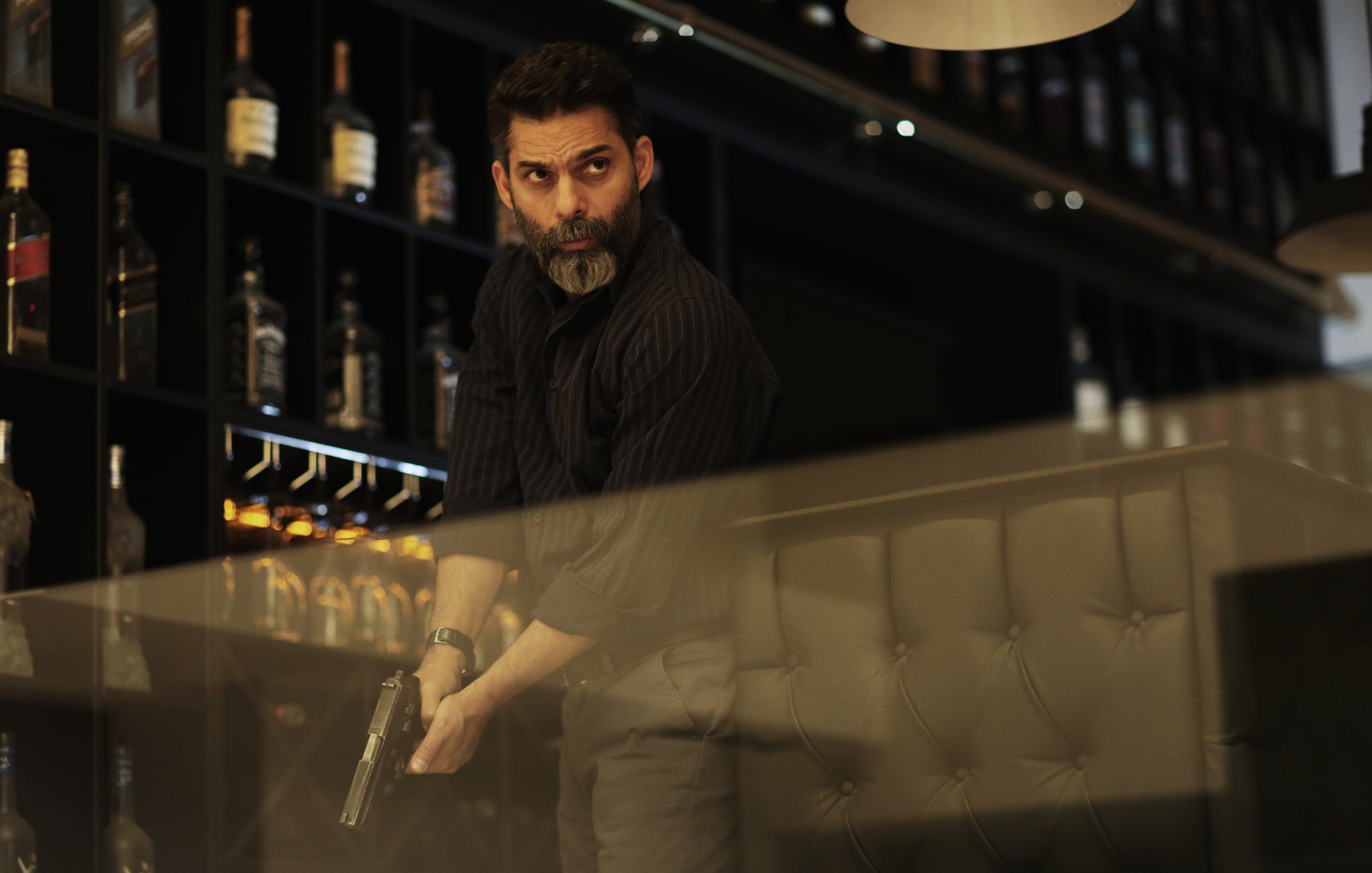 L'image montre un homme, probablement dans un bar ou un restaurant, tenant une arme à feu. Il a une barbe grise et une expression sérieuse, suggérant qu'il est dans une situation tendue. Le décor en arrière-plan comprend des étagères avec des bouteilles de spiritueux. L'éclairage est tamisé, créant une atmosphère dramatique.