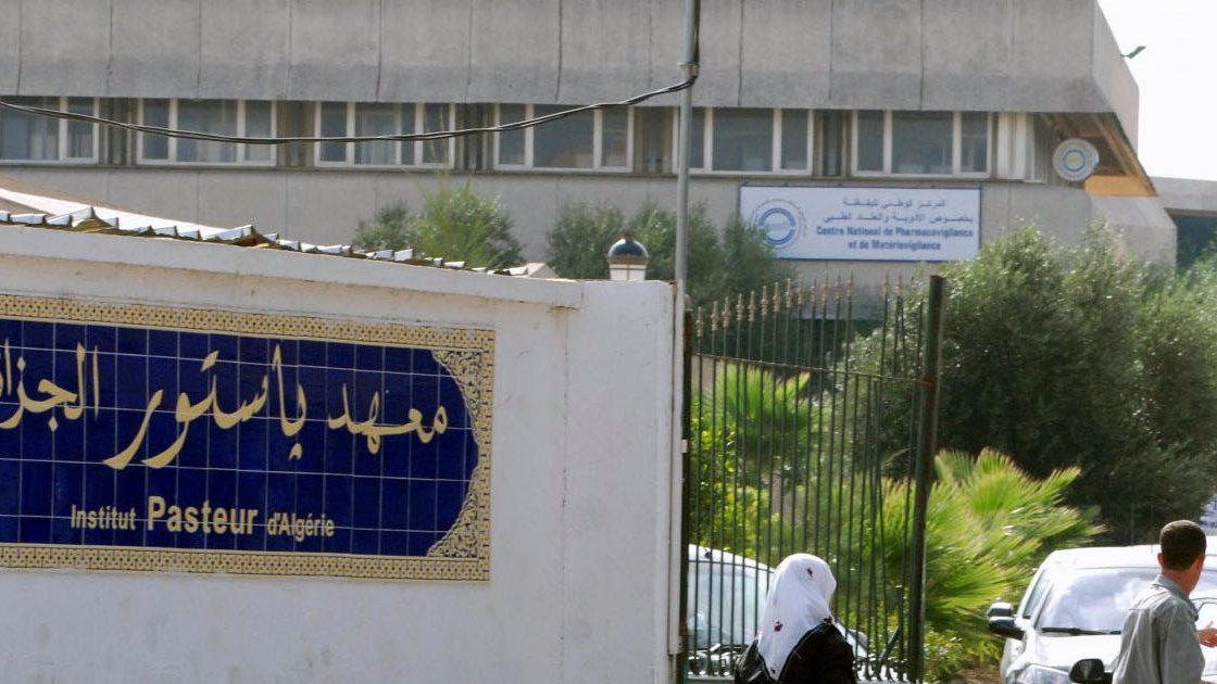 L'image montre l'extérieur de l'Institut Pasteur en Algérie. On peut voir un bâtiment moderne en arrière-plan avec des panneaux d'affichage. À l'avant-plan, il y a une entrée avec une façade blanche où figure le nom de l'institut en caractères arabes et français. Quelques personnes marchent dans cette zone, suggérant un environnement fréquenté. Des arbres et de la végétation ajoutent une touche de verdure au décor.