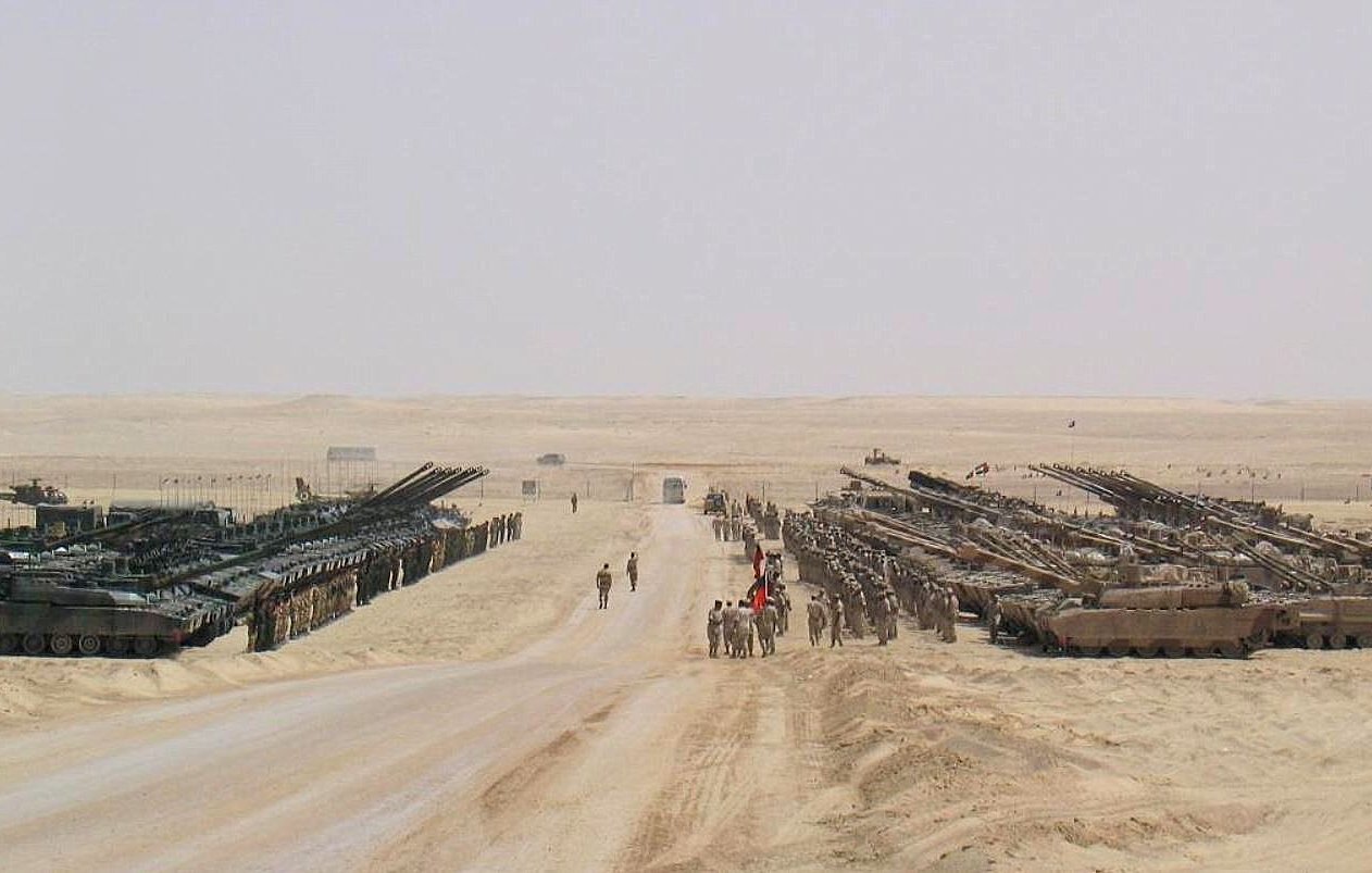 L'image montre un paysage désertique avec des formations militaires. On y voit des chars d'assaut alignés de chaque côté d'une route en terre, tandis que plusieurs soldats sont présents sur le chemin. Le terrain est aride et poussiéreux, avec un ciel clair et peu de végétation. L'atmosphère dégage une impression de préparation militaire ou d'exercice.
