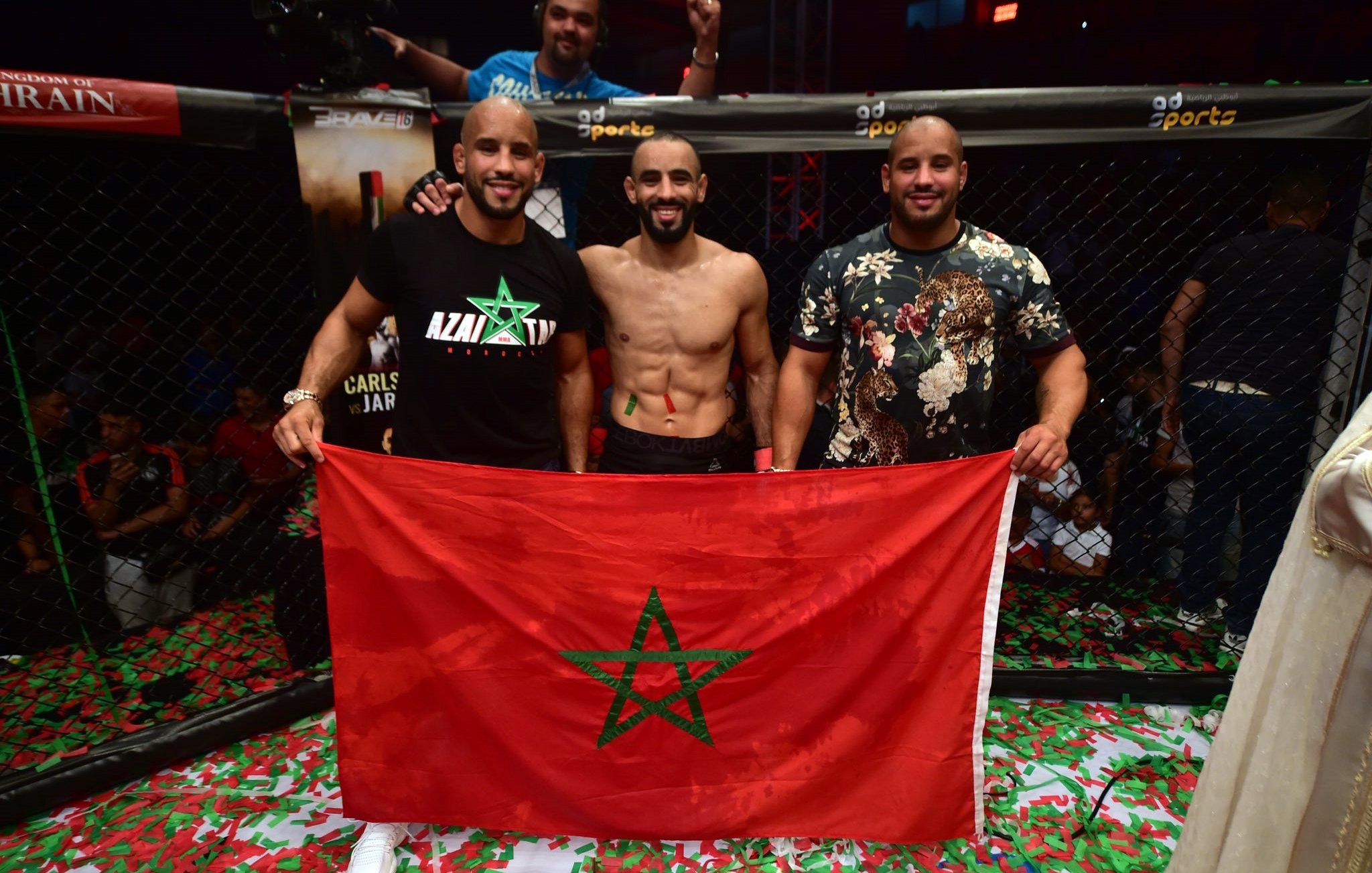 L'image montre trois hommes posant ensemble à l'intérieur d'un environnement de combat, probablement après une victoire. L'un d'eux est torse nu et affiche un large sourire, tandis que les autres sont habillés avec des t-shirts. Ils tiennent une grande drapeau marocain, mettant en avant leur fierté nationale. Le sol est couvert de confettis, ajoutant une atmosphère festive à la scène. En arrière-plan, on peut voir des spectateurs soulevant les bras en signe de célébration.