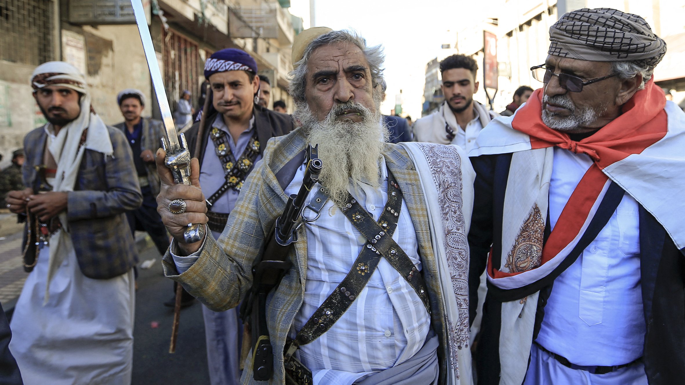 L'image montre un groupe d'hommes marchant dans une rue. L'un d'eux, au centre, est une personne âgée avec une longue barbe grise, tenant une épée. Il porte une tenue traditionnelle composée d'une chemise et d'une veste, ainsi qu'une ceinture. Les autres hommes dans le groupe portent aussi des vêtements traditionnels, et certains semblent porter des accessoires tels que des médaillons ou des fusils. L'ambiance de l'image suggère une manifestation ou une célébration culturelle. L'arrière-plan montre des bâtiments urbains typiques.