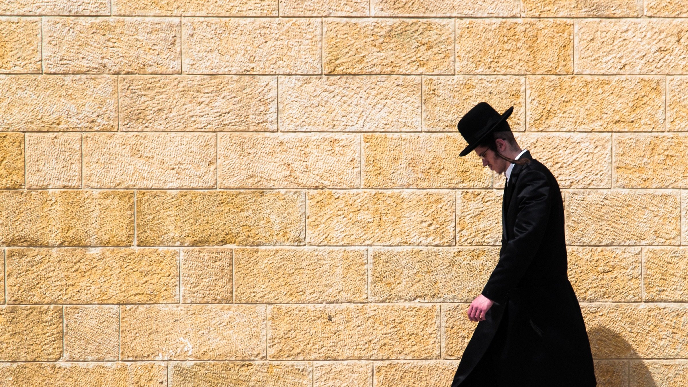L'image montre un homme marchant le long d'un mur de pierre de couleur beige. Il est habillé d'un long manteau noir et porte un chapeau noir. Son expression est sérieuse et il semble concentré sur son chemin. L'arrière-plan est uni, ce qui met en valeur la silhouette de l'homme qui avance seul.