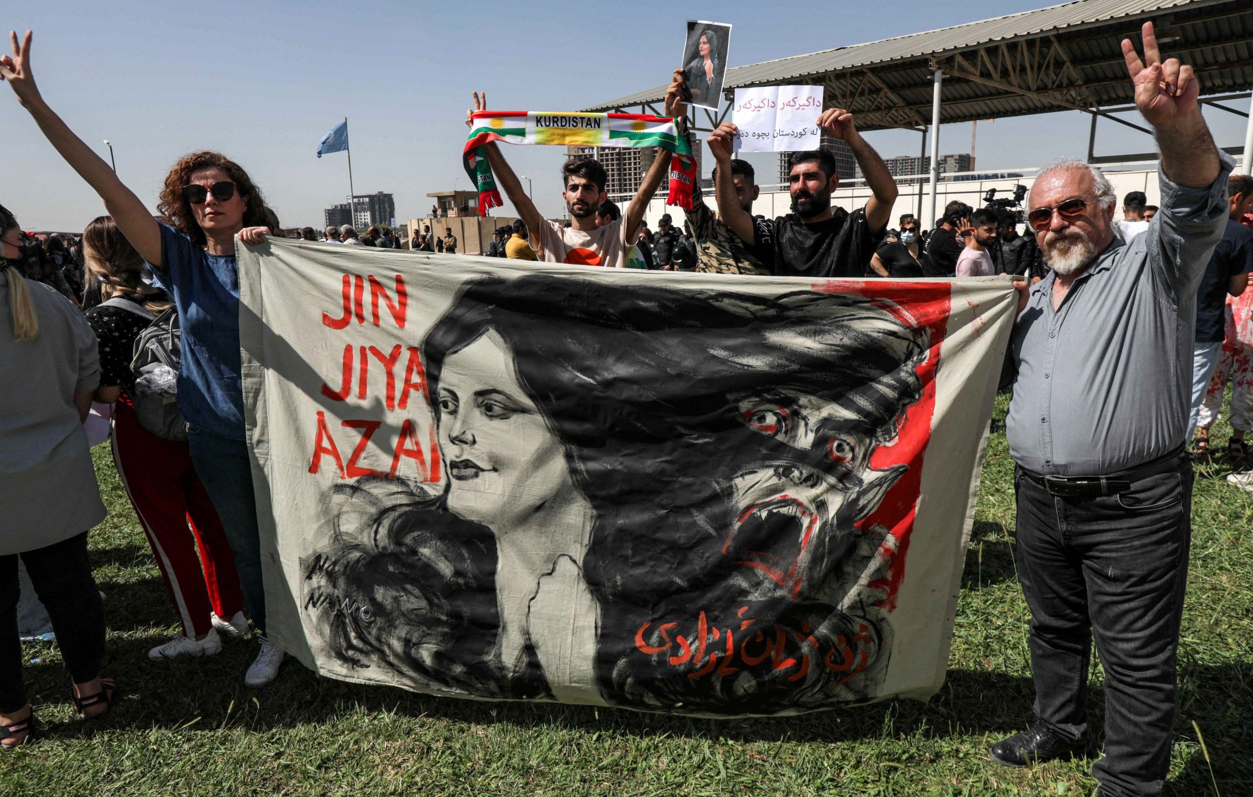 L'image montre une scène de manifestation. Des personnes tiennent une grande bannière sur laquelle est représentée le visage d'une femme entouré de motifs dramatiques. Les manifestants affichent des signes de paix avec leurs mains. On peut voir des drapeaux, probablement kurdes, en arrière-plan. L'atmosphère semble chargée d'émotion et de détermination, suggérant un rassemblement pour une cause importante.