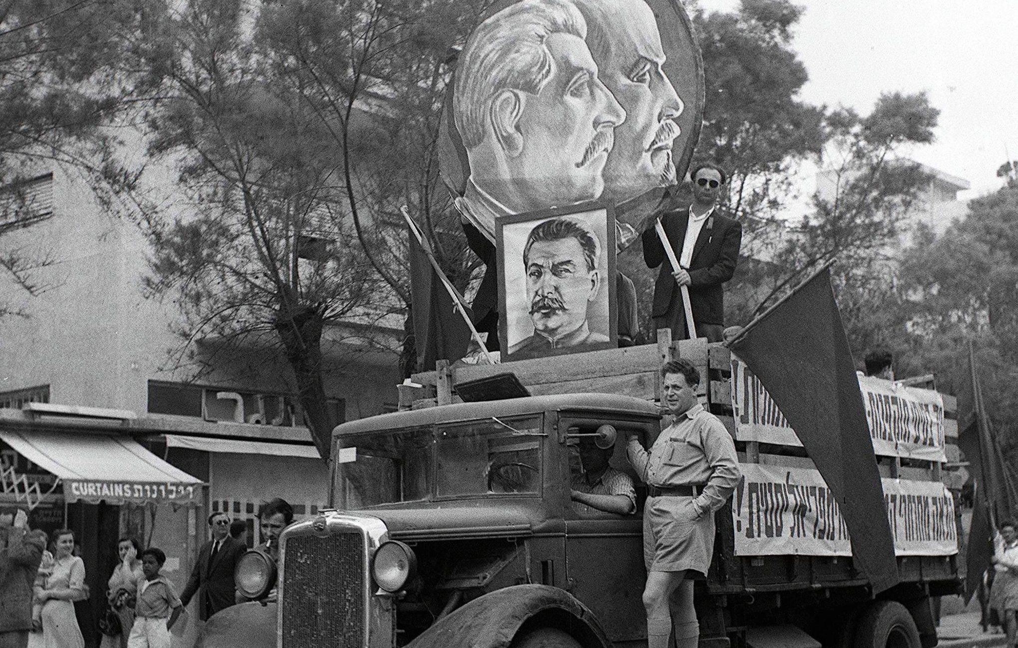 L'image représente un défilé historique avec un camion décoré. Sur le camion, on peut voir des portraits de figures politiques, probablement des leaders communistes, entourés de drapeaux. Un homme en uniforme se tient debout sur le camion, tandis que d'autres personnes défilent en arrière-plan. L'atmosphère semble festive et engagée, typique des manifestations politiques d'époque. Les bâtiments et les passants en arrière-plan ajoutent un contexte urbain à la scène.