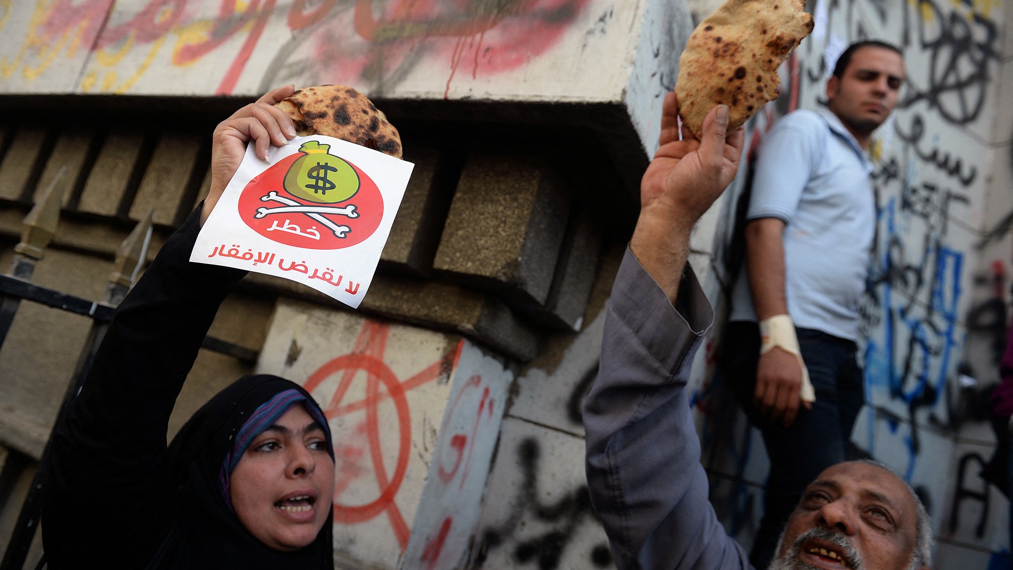 L'image montre des manifestants tenant des pains et des pancartes. Une femme en hijab et un homme plus âgé semblent exprimer leur mécontentement. En arrière-plan, on peut voir des murs couverts de graffitis, et une autre personne se tient debout. L'atmosphère semble chargée d'émotion, soulignant une protestation ou une revendication sociale.