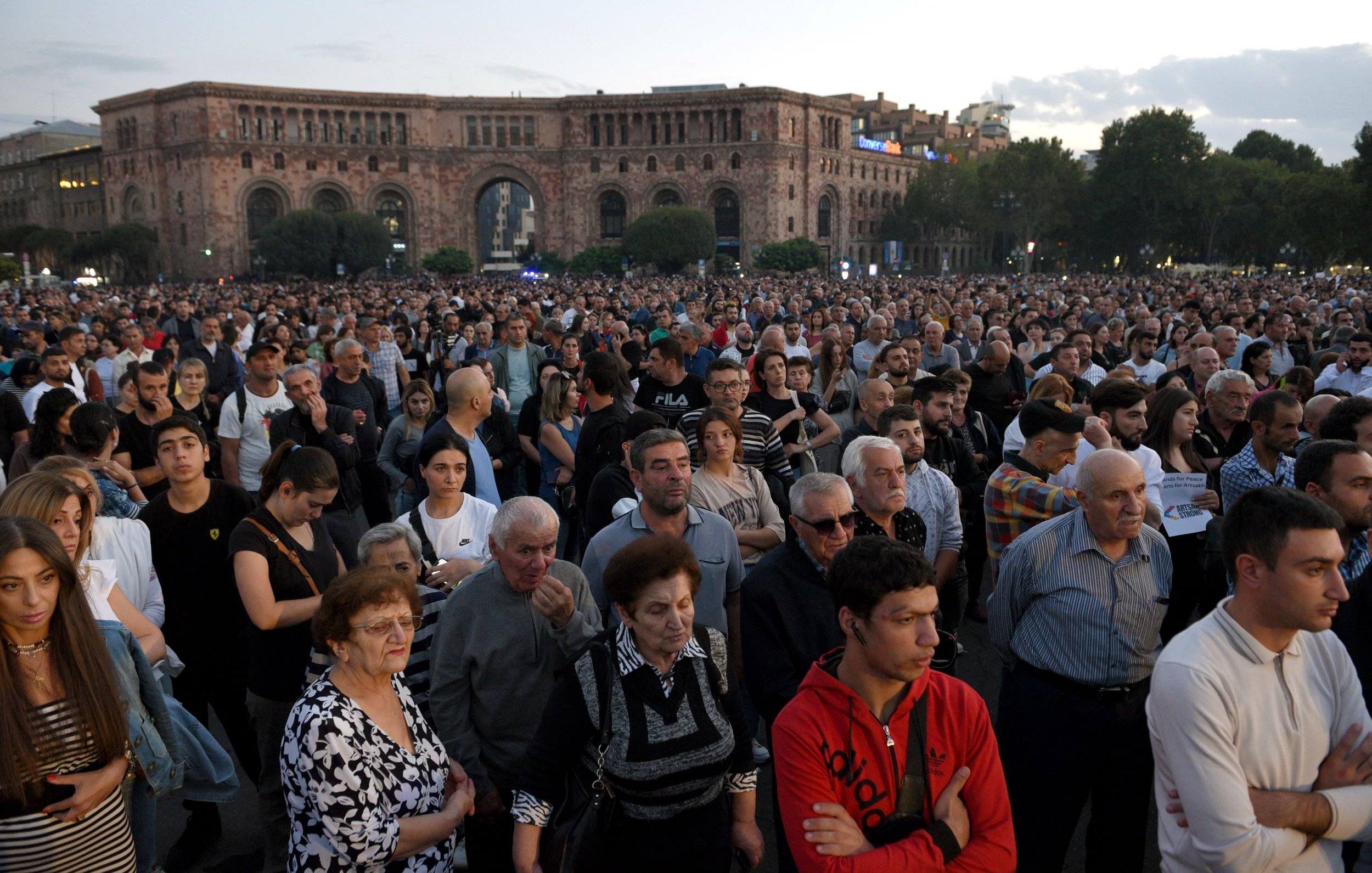 L'image montre une grande foule de personnes rassemblées dans un espace public, probablement pour un événement ou une manifestation. Les gens semblent concentrés et attentifs, certains regardant vers l'avant. L'arrière-plan présente des bâtiments, suggérant un milieu urbain, et le ciel est légèrement nuageux, indiquant que le jour commence à tomber. L'atmosphère semble à la fois solennelle et engagée.