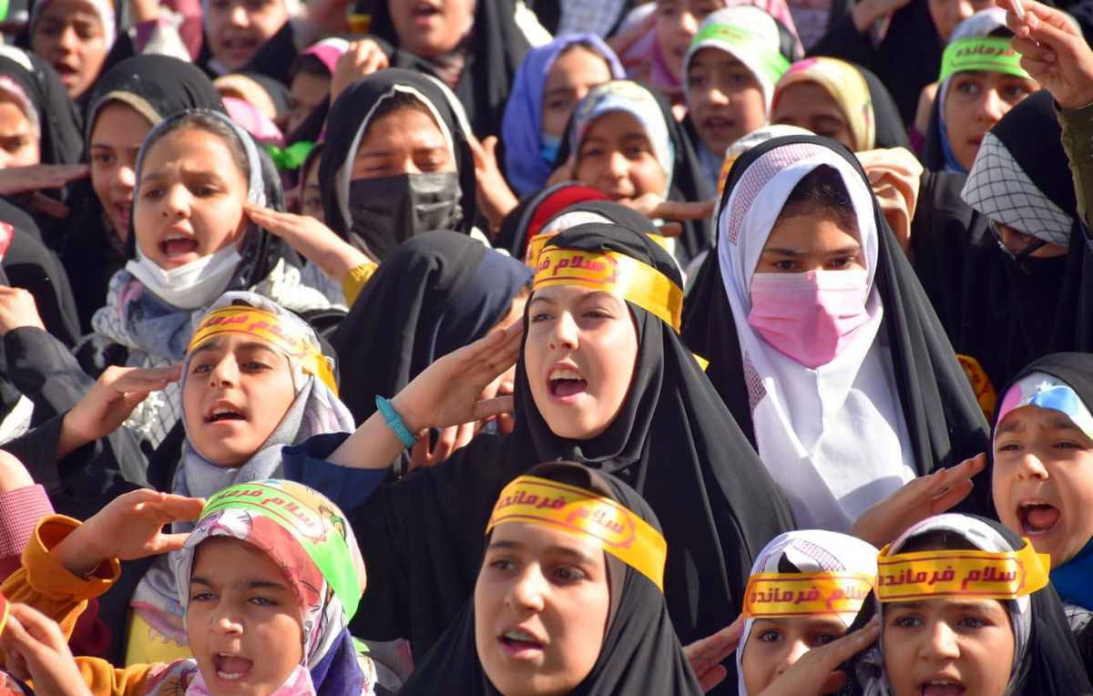 L'image montre un grand groupe de jeunes filles, portant des hijabs et des bandeaux avec des inscriptions en arabe. Elles semblent exprimer de l'enthousiasme, chantant ou criant, et leurs visages sont pleins d'énergie. L'arrière-plan indique un rassemblement, probablement lié à un événement spécial ou une manifestation. Les couleurs des vêtements et des bandeaux apportent une atmosphère vibrante à la scène.