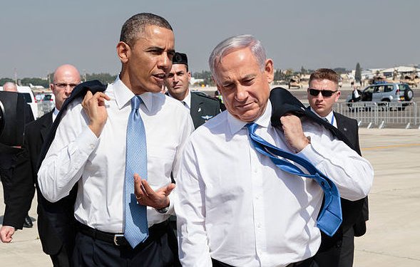 L'image montre deux hommes, habillés en costume, marchant ensemble sur un tarmac. L'un porte une cravate bleue et l'autre une cravate claire. Ils semblent engagés dans une conversation. En arrière-plan, on peut apercevoir des membres de la sécurité et des véhicules. Le ciel est clair, et l'environnement semble être un aéroport.